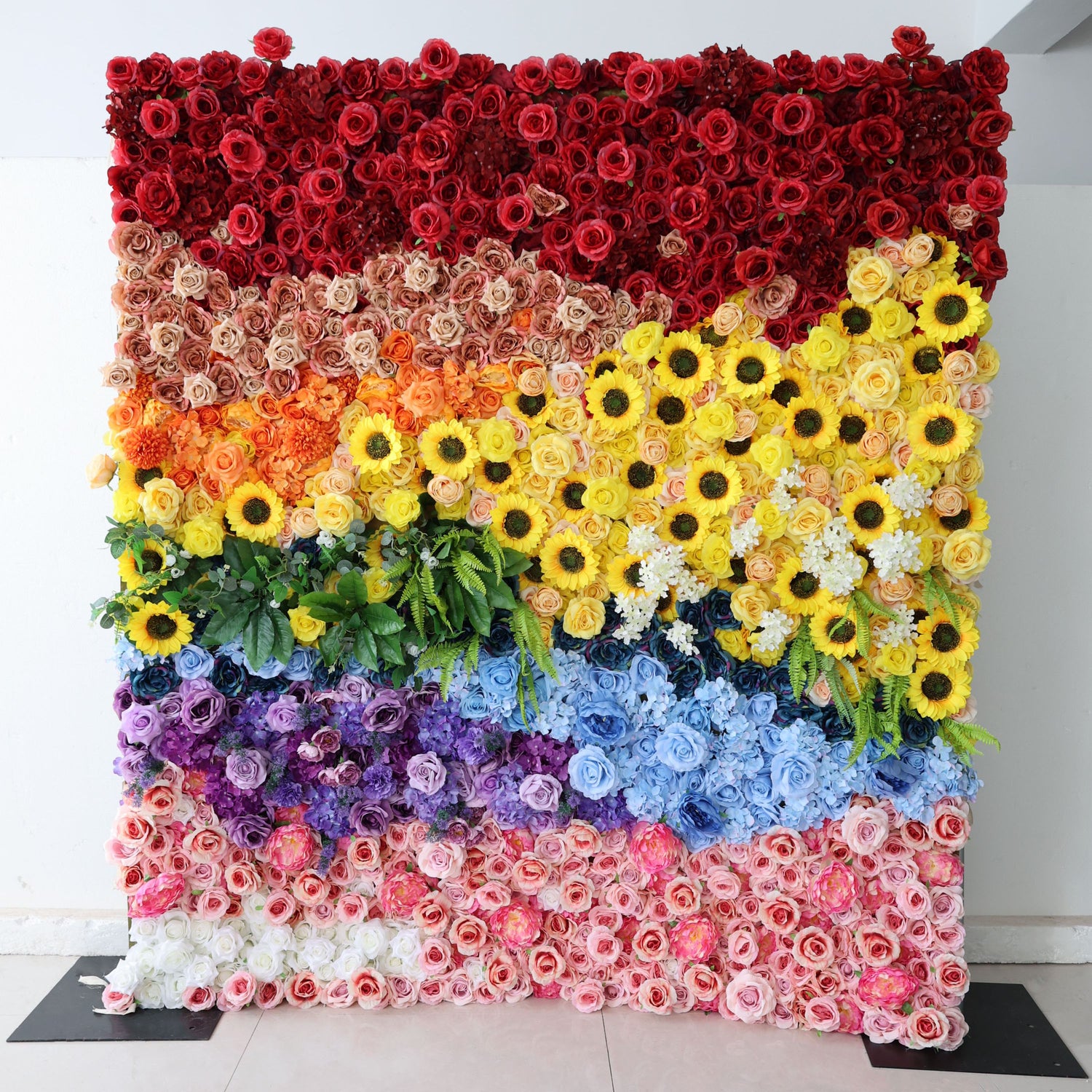 This image showcases a vibrant floral wall by Valar Flowers. It features a stunning arrangement of artificial flowers in a rainbow of colors—deep reds, soft creams, bright yellows with sunflowers, various blues, purples, and pinks—interspersed with lush green foliage. The layered, colorful design creates a lively and eye-catching display, perfect for adding a touch of natural beauty and charm to events or interior spaces.