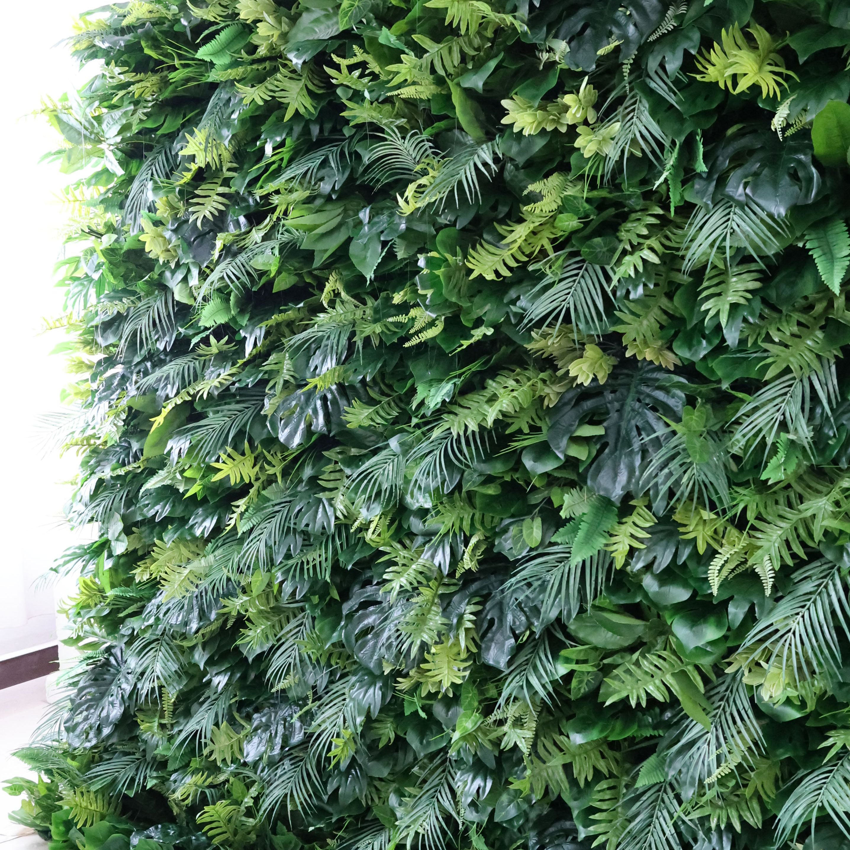 This is a dense, textured greenery panel (likely a decor piece) brimming with layered foliage: varied ferns, tropical fronds, and broad leaves in gradients of deep green and soft lime. Soft, directional light (from the left) highlights the lush, organic texture, casting subtle shadows that enhance depth. The piece feels vibrant and earthy—ideal as an event backdrop or accent wall, blending natural texture with a fresh, versatile aesthetic suited for indoor spaces.