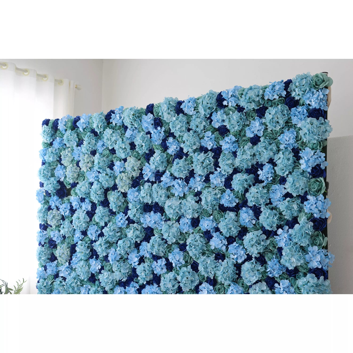 A large artificial flower wall showcases dense, layered blue hydrangeas in soft and deep tones, interspersed with green foliage. The rich, harmonious color palette creates a serene, elegant backdrop, perfect for adding a touch of refined beauty to events or decor.