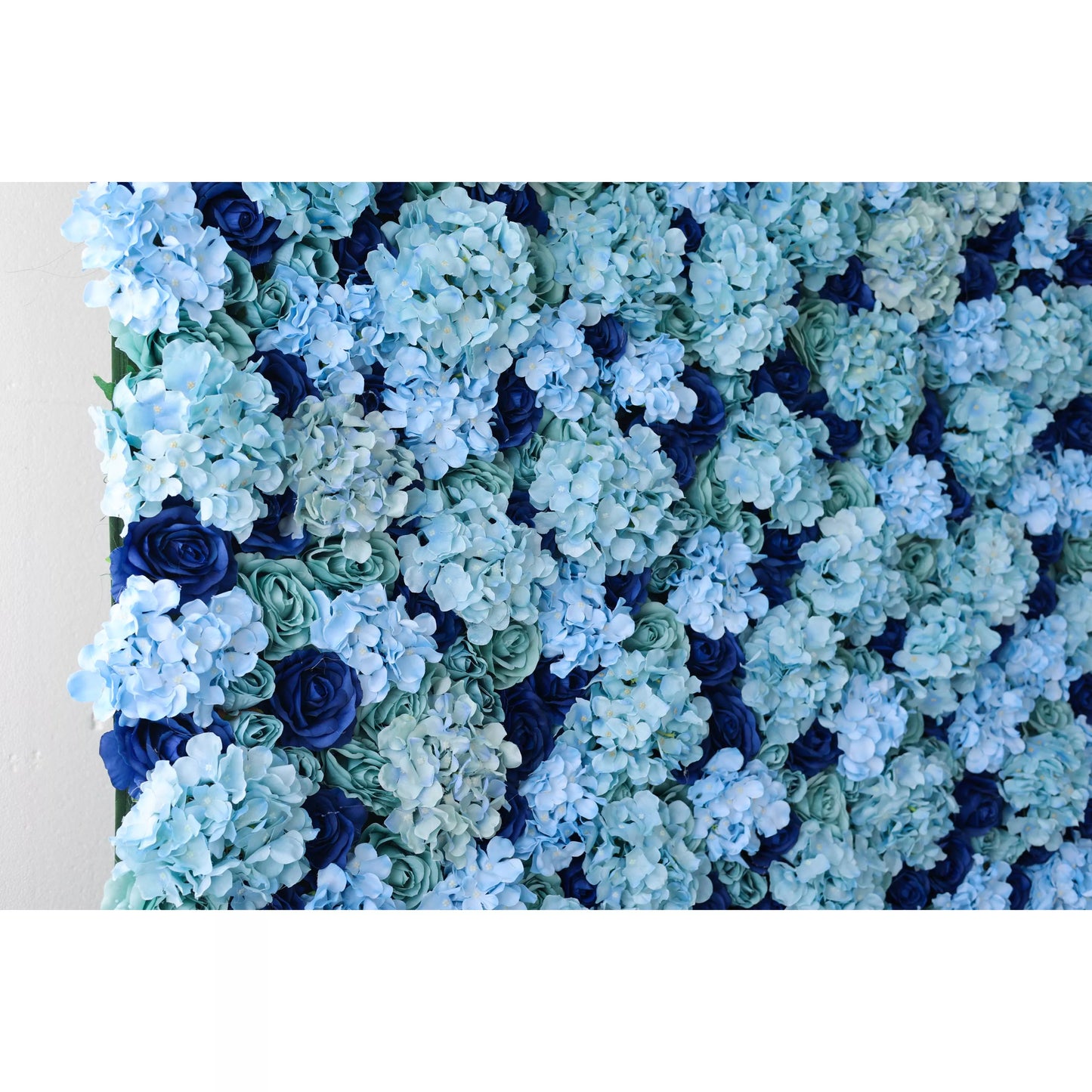 This close - up shows a lush artificial flower wall with soft blue hydrangeas, dark blue roses, and teal foliage. The layered blooms create a rich, textured, and elegant display, perfect for adding a refined, decorative touch to events or spaces.