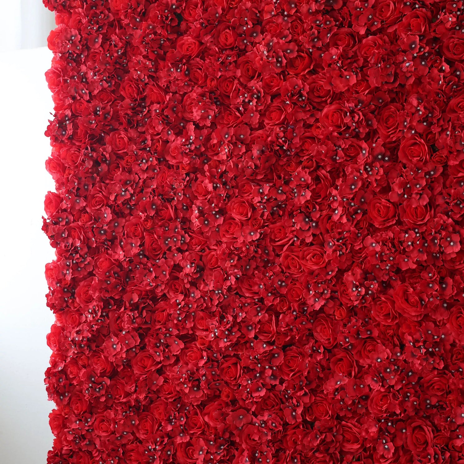 This is a large artificial flower wall, densely packed with vibrant red roses and small blossoms. The uniform, lush arrangement creates a bold, passionate, and elegant visual. It serves as a striking backdrop, perfect for adding drama and romance to events like weddings, photo shoots, or special celebrations, enhancing the atmosphere with its rich color and intricate floral design.