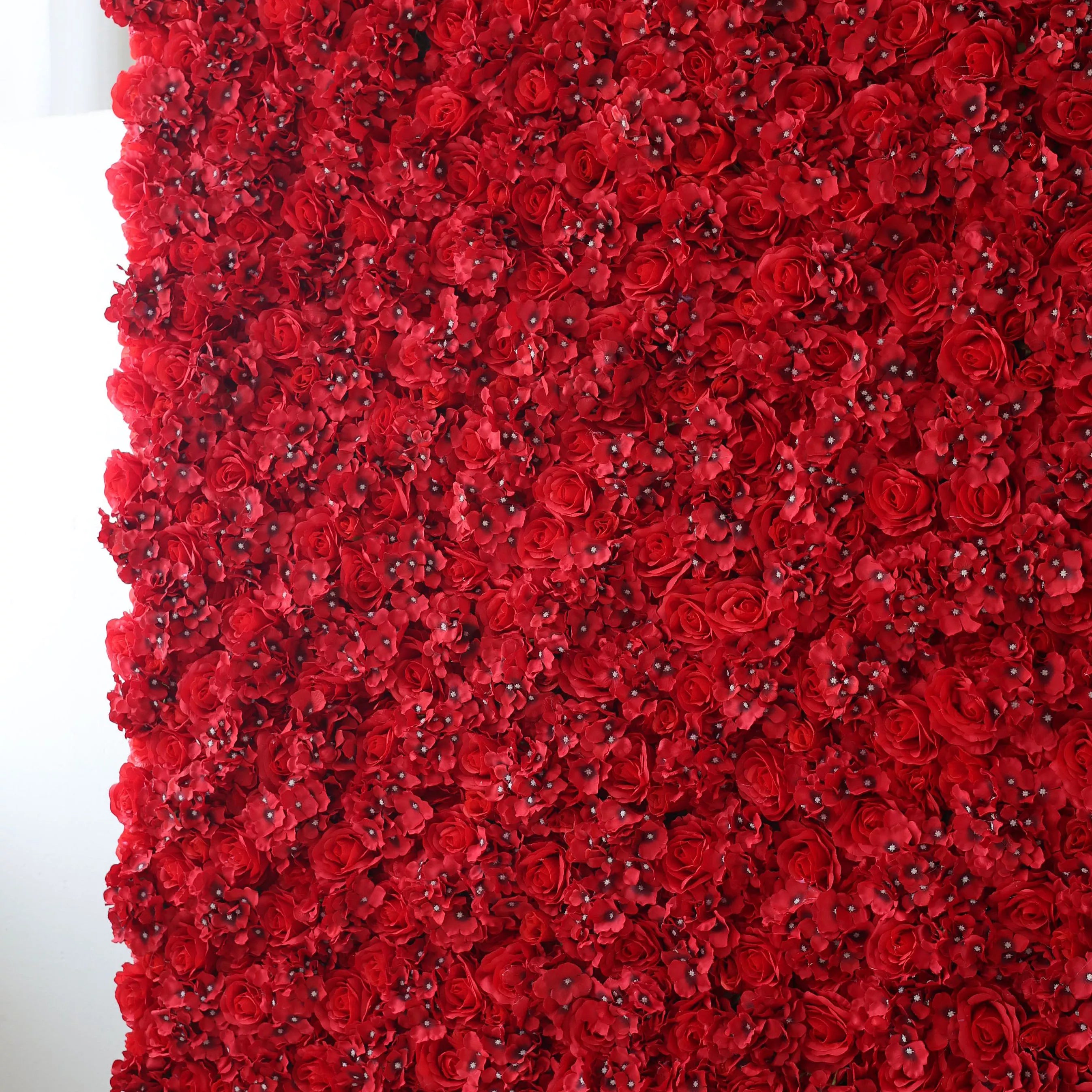 This is a large artificial flower wall, densely packed with vibrant red roses and small blossoms. The uniform, lush arrangement creates a bold, passionate, and elegant visual. It serves as a striking backdrop, perfect for adding drama and romance to events like weddings, photo shoots, or special celebrations, enhancing the atmosphere with its rich color and intricate floral design.