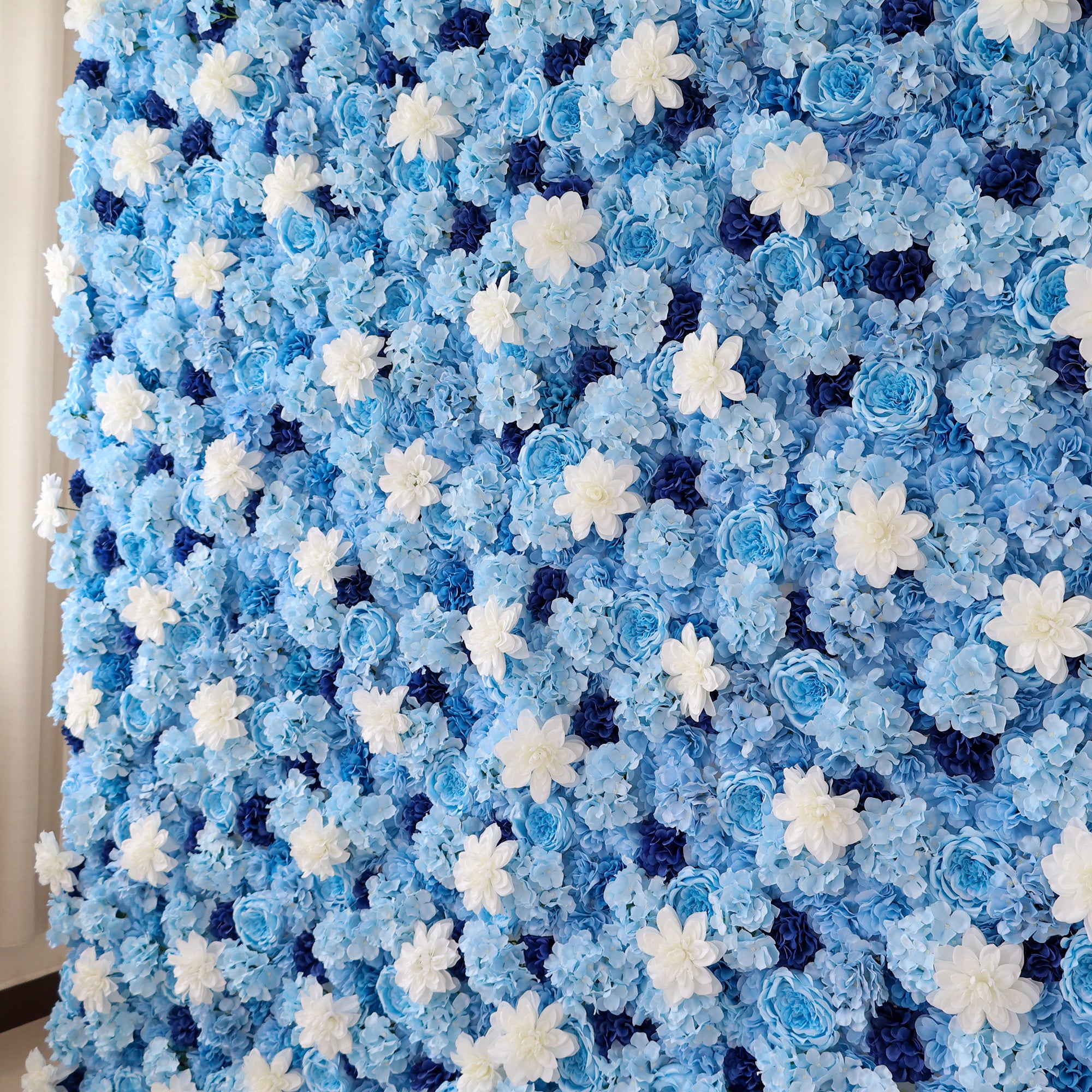 A close - up of a large artificial flower wall features blue hydrangeas, white daisy - like blooms, and dark blue blossoms. The dense, uniform arrangement creates a serene, elegant backdrop, perfect for events, evoking a calm and sophisticated vibe.
