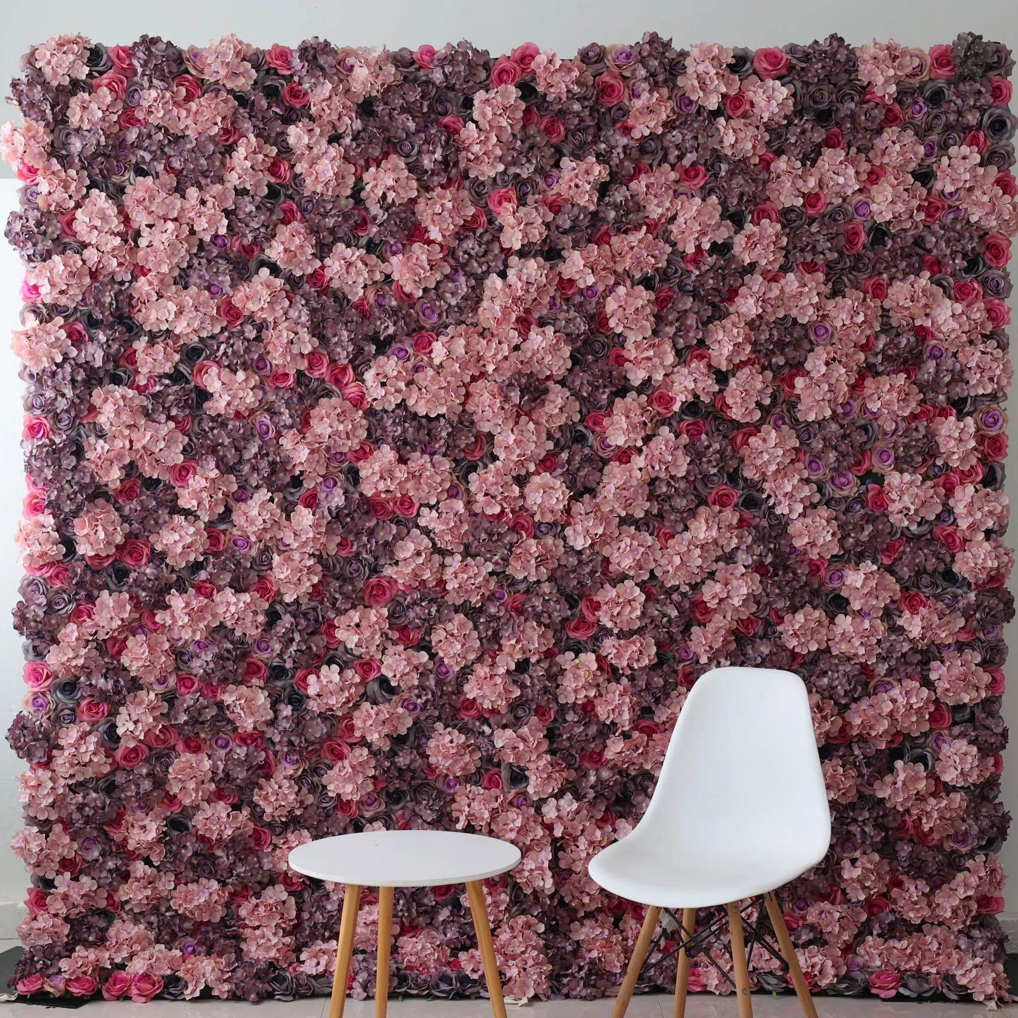This picture showcases a large artificial flower wall, a stunning blend of pink hydrangeas, purple blooms, and red roses, creating a lush, romantic backdrop. In front, a minimalist white chair and a small round table with wooden legs offer a modern, contrasting element. The combination of elaborate floral decor and simple furniture makes it an ideal setup for stylish photo shoots or elegant event spaces, exuding an atmosphere of refined beauty.