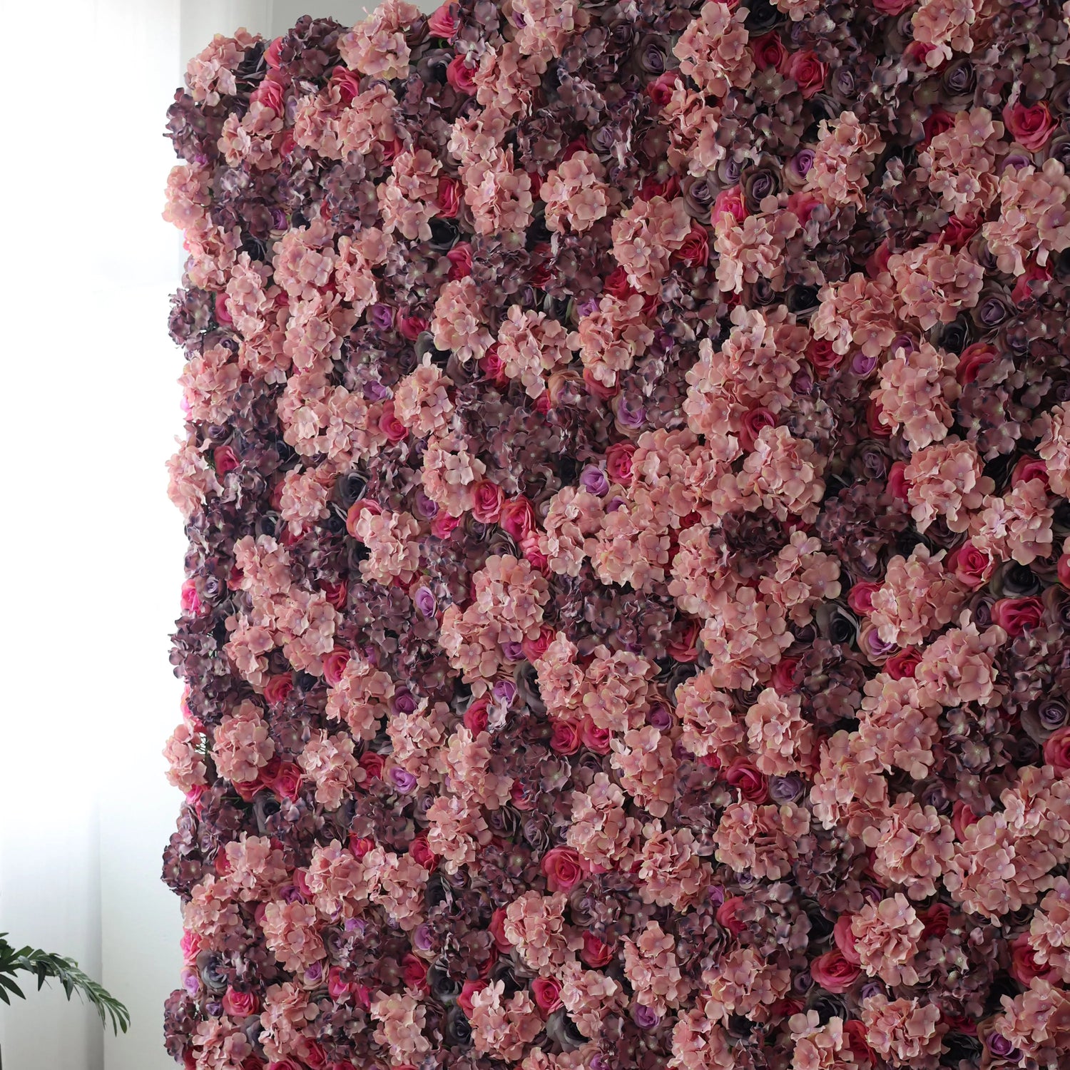 This is a large artificial flower wall, a captivating blend of pink hydrangeas, purple blooms, and red roses. The dense, layered arrangement creates a lush, romantic texture, exuding an air of elegance. Ideal for photo backdrops or event decor, it infuses spaces with refined beauty, making every moment feel special and visually enchanting.