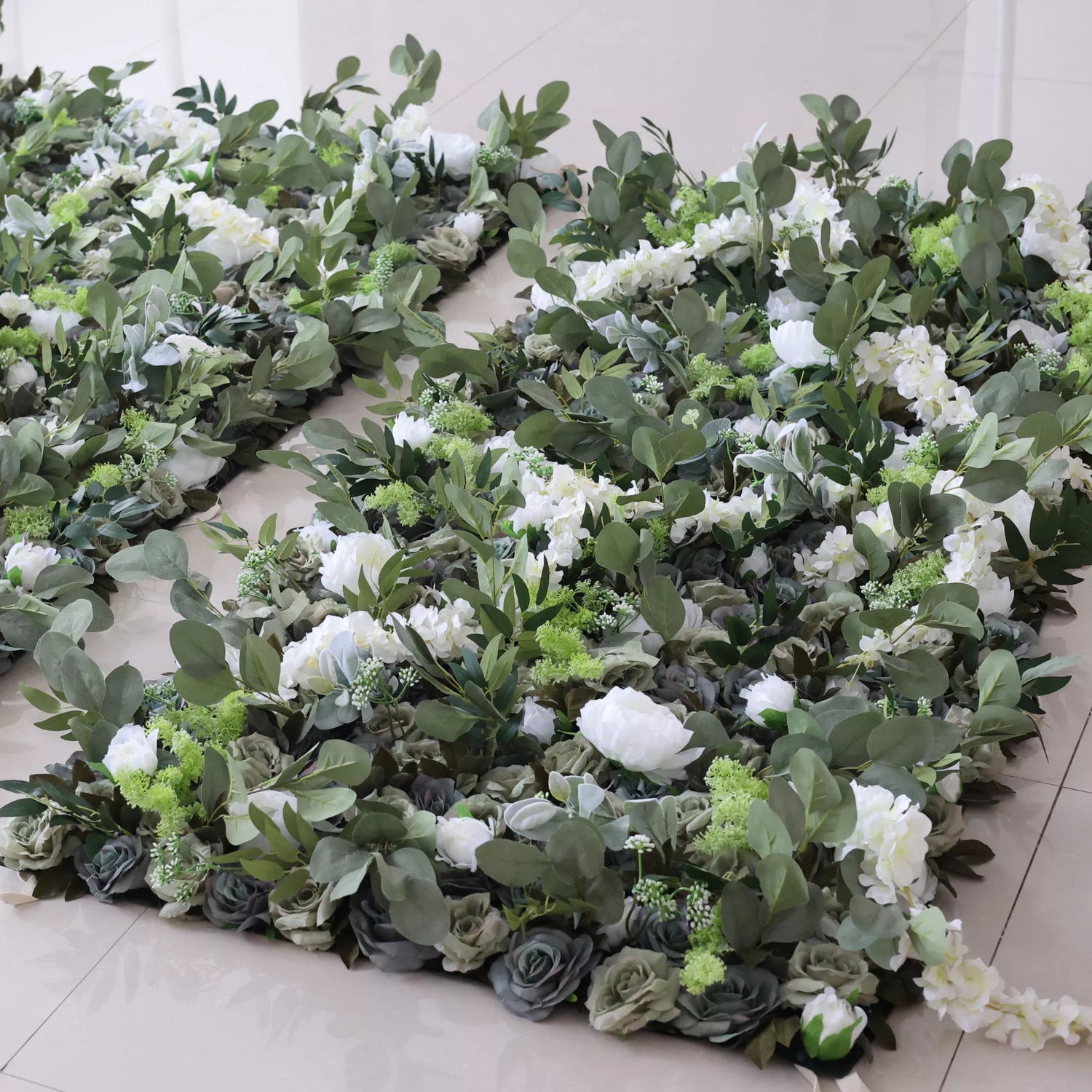 This picture shows several pieces of artificial floral decor. They feature a mix of white blooms, green foliage, and gray roses, arranged on a light - colored tiled floor. A small blue circle is visible in the center, adding a distinct detail. The lush, layered design creates an elegant, natural - looking display, perfect for decorative use.