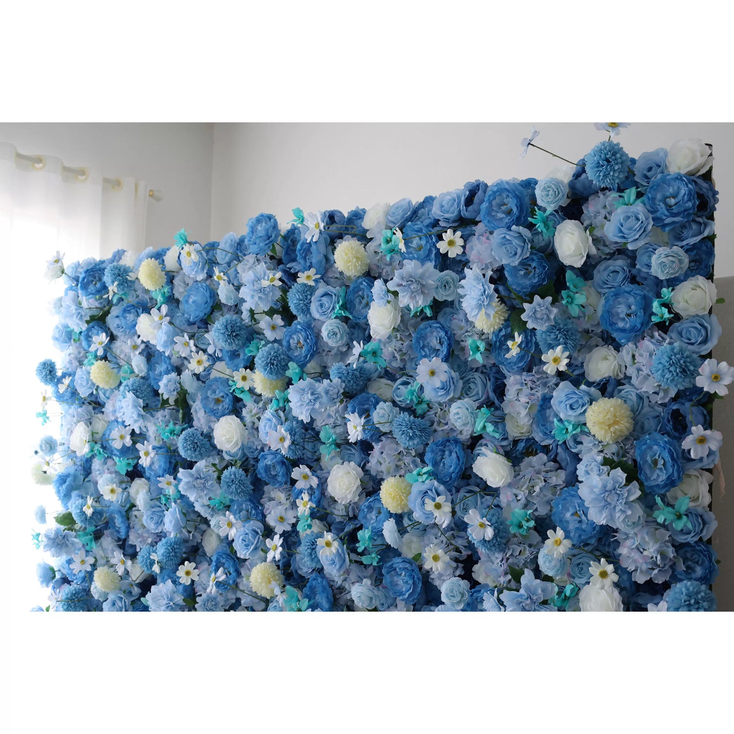 A large artificial flower wall showcases a lush blend of blue blooms, including roses, hydrangeas, and delicate white flowers with yellow centers. Accents of soft - yellow pom - pom flowers and teal foliage add texture. The harmonious mix of shades creates a serene, elegant backdrop, perfect for adding a touch of beauty to events or decor.