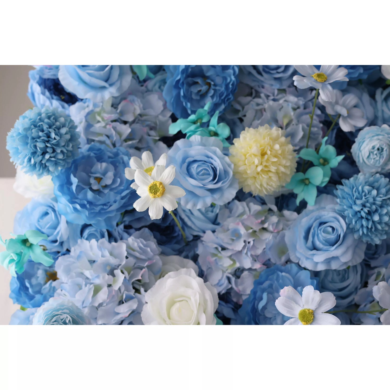 A close - up of an artificial flower wall showcases a beautiful blend of blue blooms. There are soft - toned roses, fluffy chrysanthemums, delicate daisies with yellow centers, and hints of teal foliage. The varied textures and harmonious blue hues create a lush, elegant and serene floral tapestry, perfect for adding a decorative touch to spaces or events.