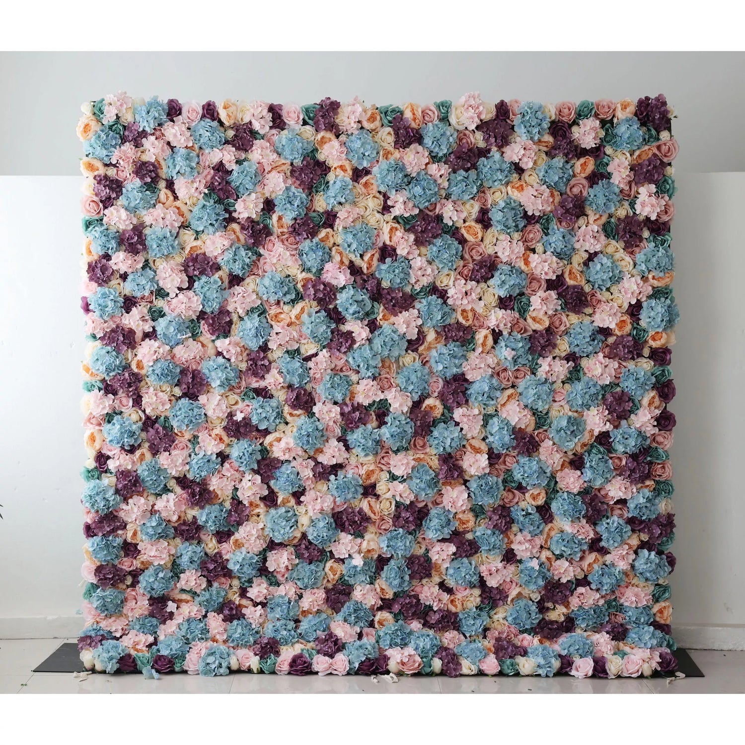This is a large artificial flower wall, showcasing a vibrant mix of colors—soft pinks, blues, purples, and creams. Densely packed with varied blooms, it creates a lush, textured tapestry. Perfect for event backdrops, adding a colorful, elegant touch to weddings or parties, enhancing the decorative appeal of any space. A small plant on the side complements the scene.
