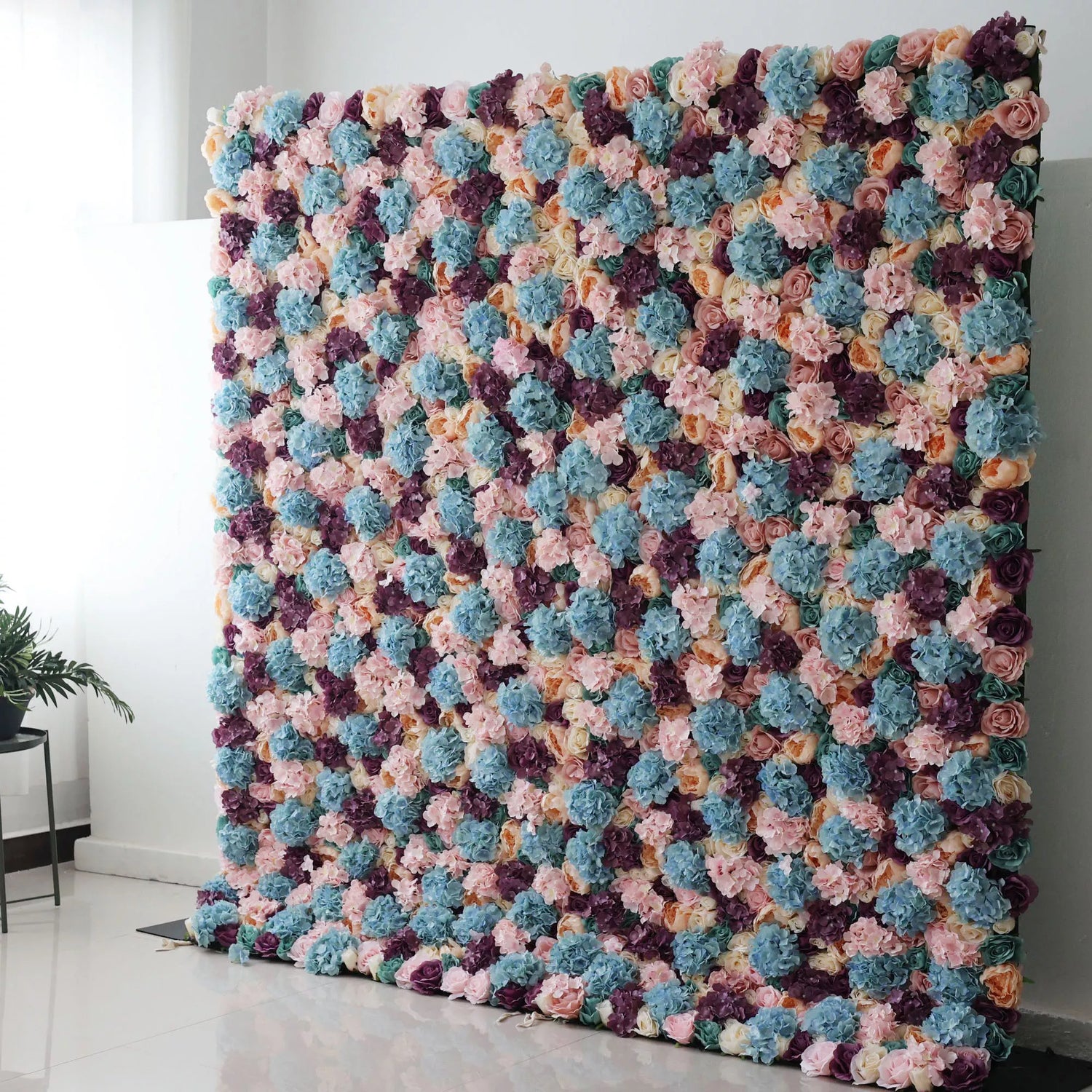 Valar Flowers Roll Up Backdrop: This is a large artificial flower wall. It showcases a vibrant mix of colors—soft pinks, blues, purples, and creams. Densely packed with varied blooms, it creates a lush, textured tapestry. Perfect for event backdrops, adding a colorful, elegant touch to weddings or parties, enhancing the decorative appeal of any space.