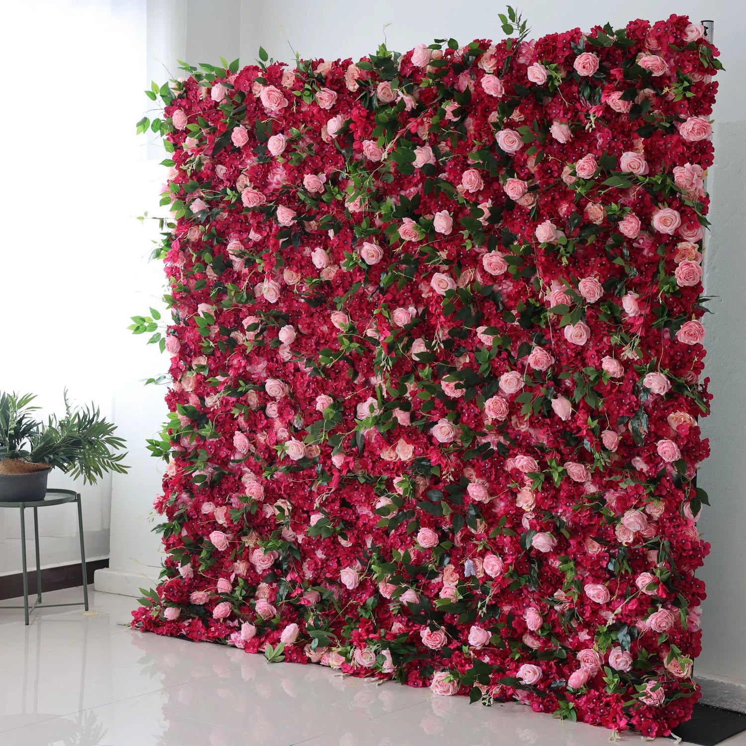 This picture features a large artificial flower wall, a vibrant mix of deep red blossoms and soft pink roses, interspersed with green leaves. The dense, lush arrangement creates a romantic and eye - catching display, perfect for event backdrops. The contrasting colors and natural - looking foliage add an elegant, decorative touch, ideal for weddings or parties to create a dreamy atmosphere. A small plant on the side complements the scene.