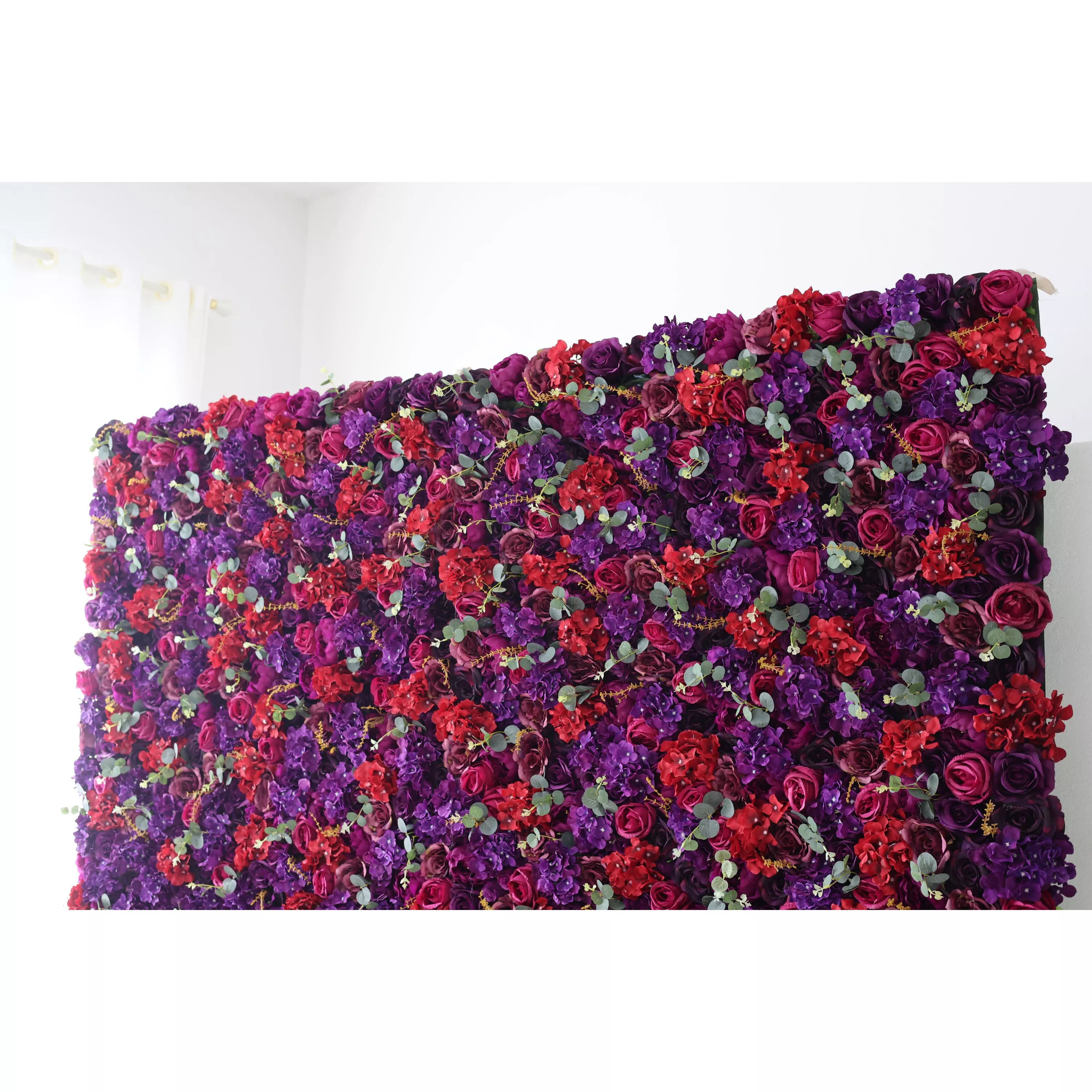 This picture presents a large artificial flower wall, a captivating mix of deep purple and vibrant red blooms. Interspersed with small green leaves, the dense arrangement creates a rich, textured tapestry. The contrasting colors and lush design make it a striking backdrop, ideal for events like weddings or parties, adding an air of elegance and drama. A bright, plain background highlights the floral display, enhancing its visual impact.