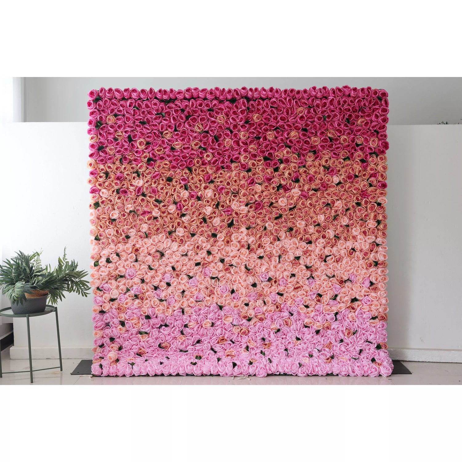 This picture shows a large artificial flower wall, a beautiful gradient of pink shades. It’s densely packed with rose - like blooms, transitioning from deep pink at the top to soft pink at the bottom. The uniform, lush arrangement creates a romantic, eye - catching display, perfect for event backdrops. A small green plant on the side adds a touch of natural contrast, enhancing the overall decorative appeal of the space.
