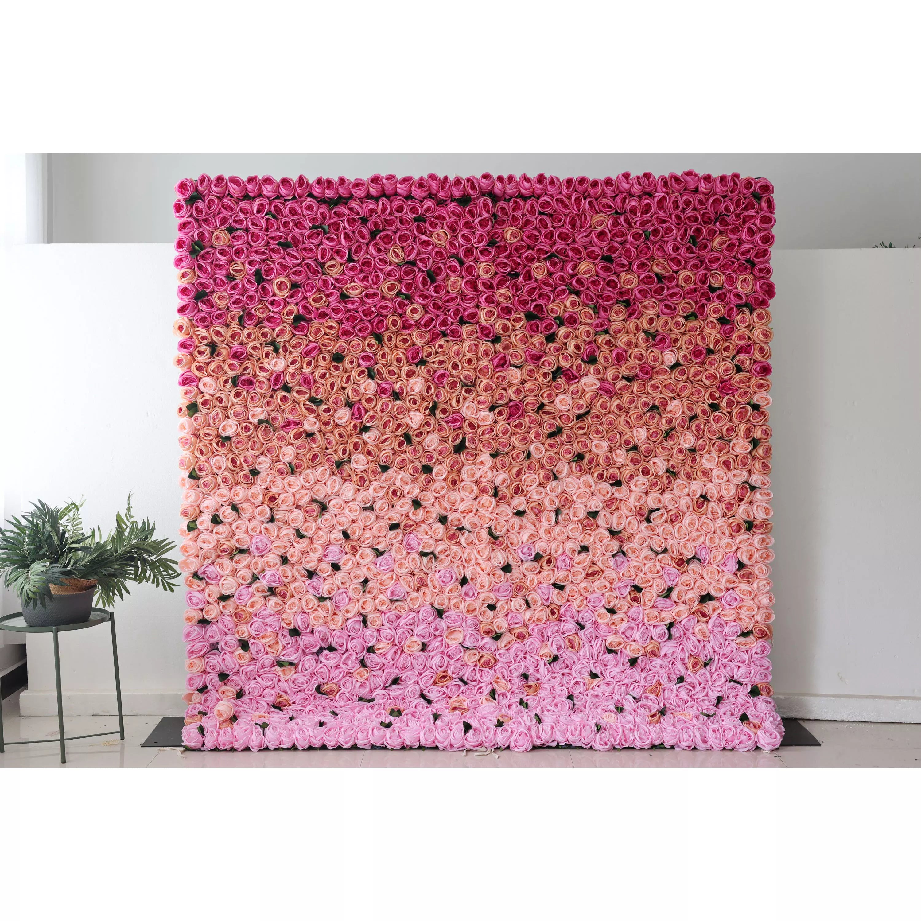 This picture shows a large artificial flower wall, a beautiful gradient of pink shades. It’s densely packed with rose - like blooms, transitioning from deep pink at the top to soft pink at the bottom. The uniform, lush arrangement creates a romantic, eye - catching display, perfect for event backdrops. A small green plant on the side adds a touch of natural contrast, enhancing the overall decorative appeal of the space.