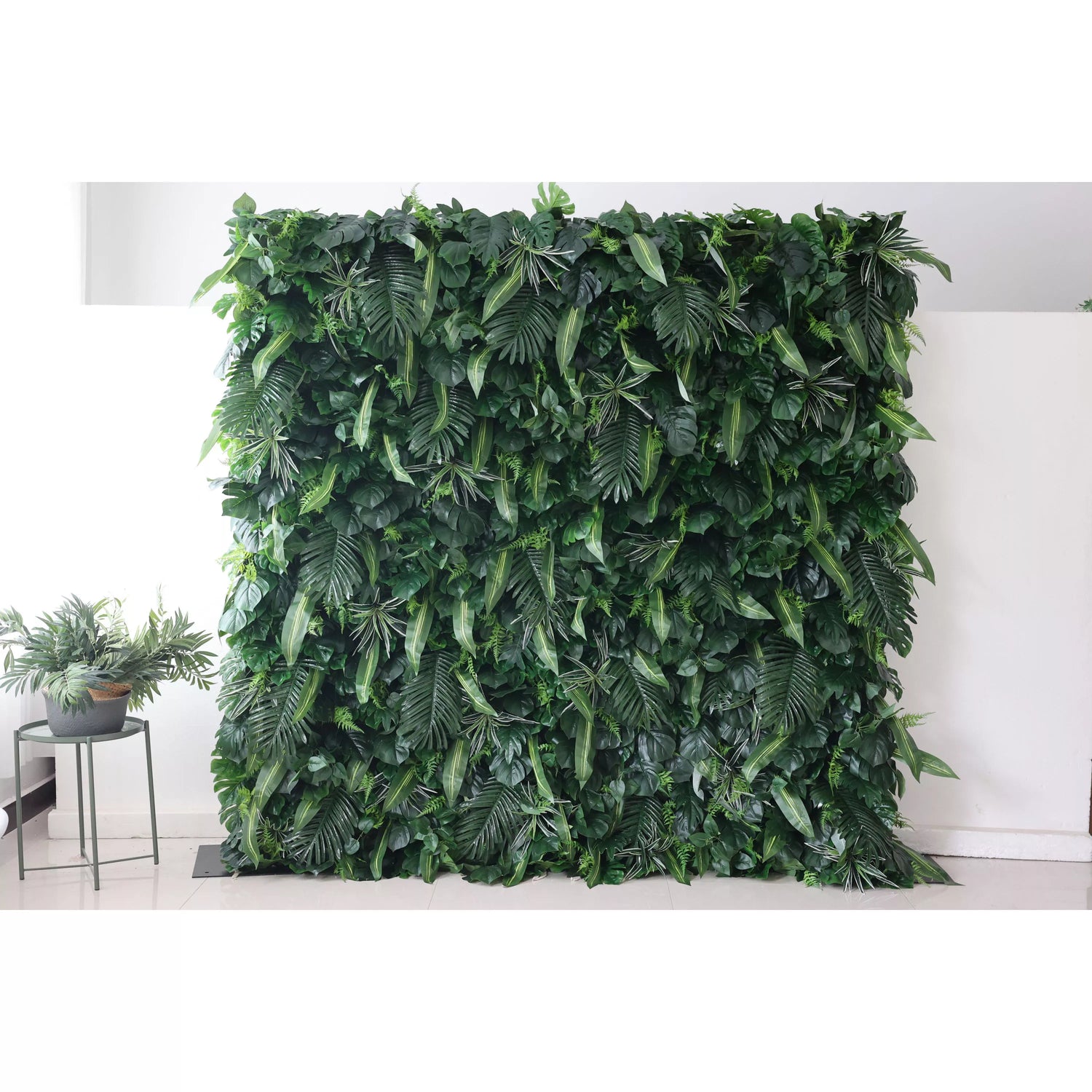 This picture presents a large, lush artificial greenery wall. It features a dense arrangement of diverse green leaves, including broad palm - like fronds and smaller, variegated foliage. The rich, verdant texture creates a natural, tropical - inspired backdrop. Placed against a plain white wall and floor, with a small plant visible on the side, it brings an instant touch of nature indoors, ideal for decorative uses in events or interior spaces.