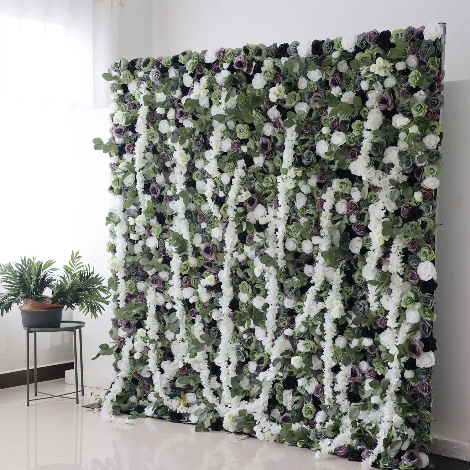This is a large artificial flower wall with a lush, green - based color scheme. It features white flowers, purple roses, and green foliage, creating a dense, natural - looking display. Paired with potted plants on the side, it adds an elegant, verdant touch, perfect for decorative event backdrops.