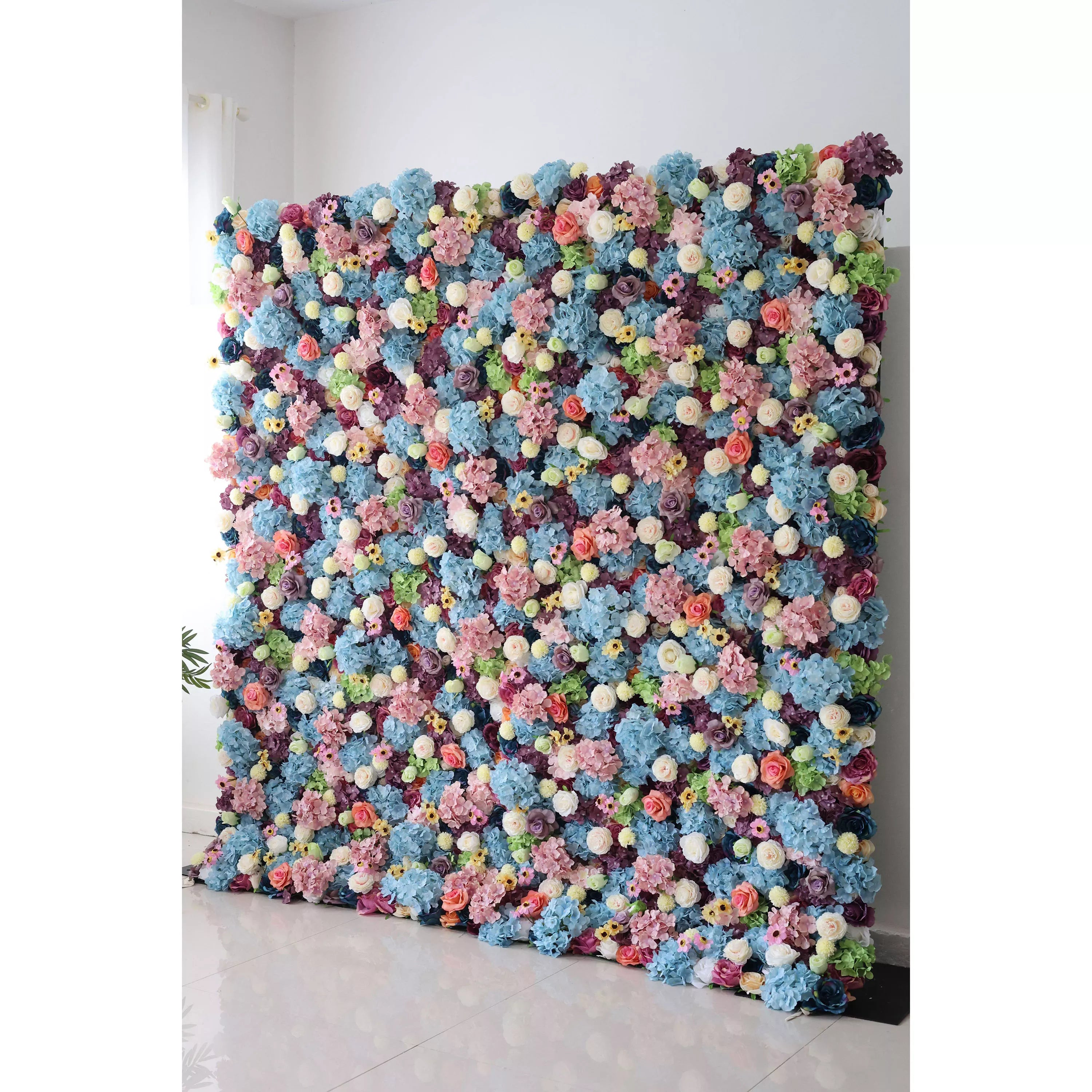 This is a large, vibrant artificial flower wall. It features a dense mix of blue hydrangeas, white roses, pink blossoms, and small yellow flowers, interspersed with green foliage. The colorful, lush arrangement creates a lively, eye - catching display, perfect for event backdrops, adding a cheerful and decorative touch.