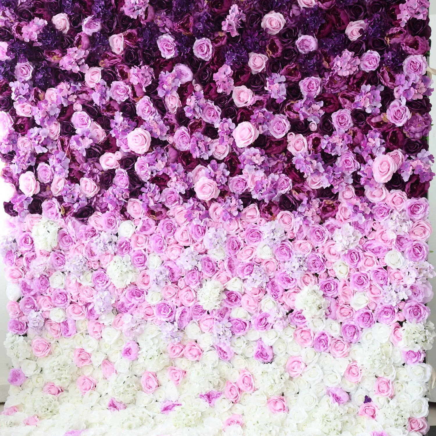 This flower wall is a stunning arrangement of artificial flowers, showcasing a gradient of colors from deep purple at the top, transitioning through various shades of pink, to white at the bottom. Comprising roses and other delicate blooms, it creates a lush, romantic texture. Ideal for weddings, parties, or other special events, it serves as an enchanting backdrop, exuding elegance and charm.