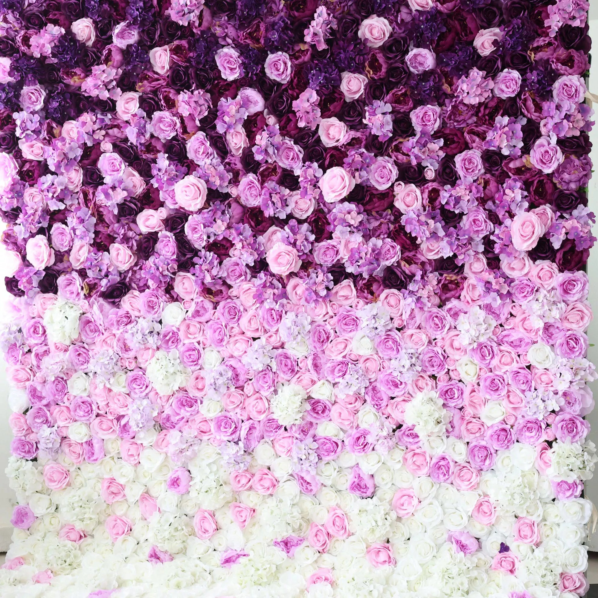 This flower wall is a stunning arrangement of artificial flowers, showcasing a gradient of colors from deep purple at the top, transitioning through various shades of pink, to white at the bottom. Comprising roses and other delicate blooms, it creates a lush, romantic texture. Ideal for weddings, parties, or other special events, it serves as an enchanting backdrop, exuding elegance and charm.
