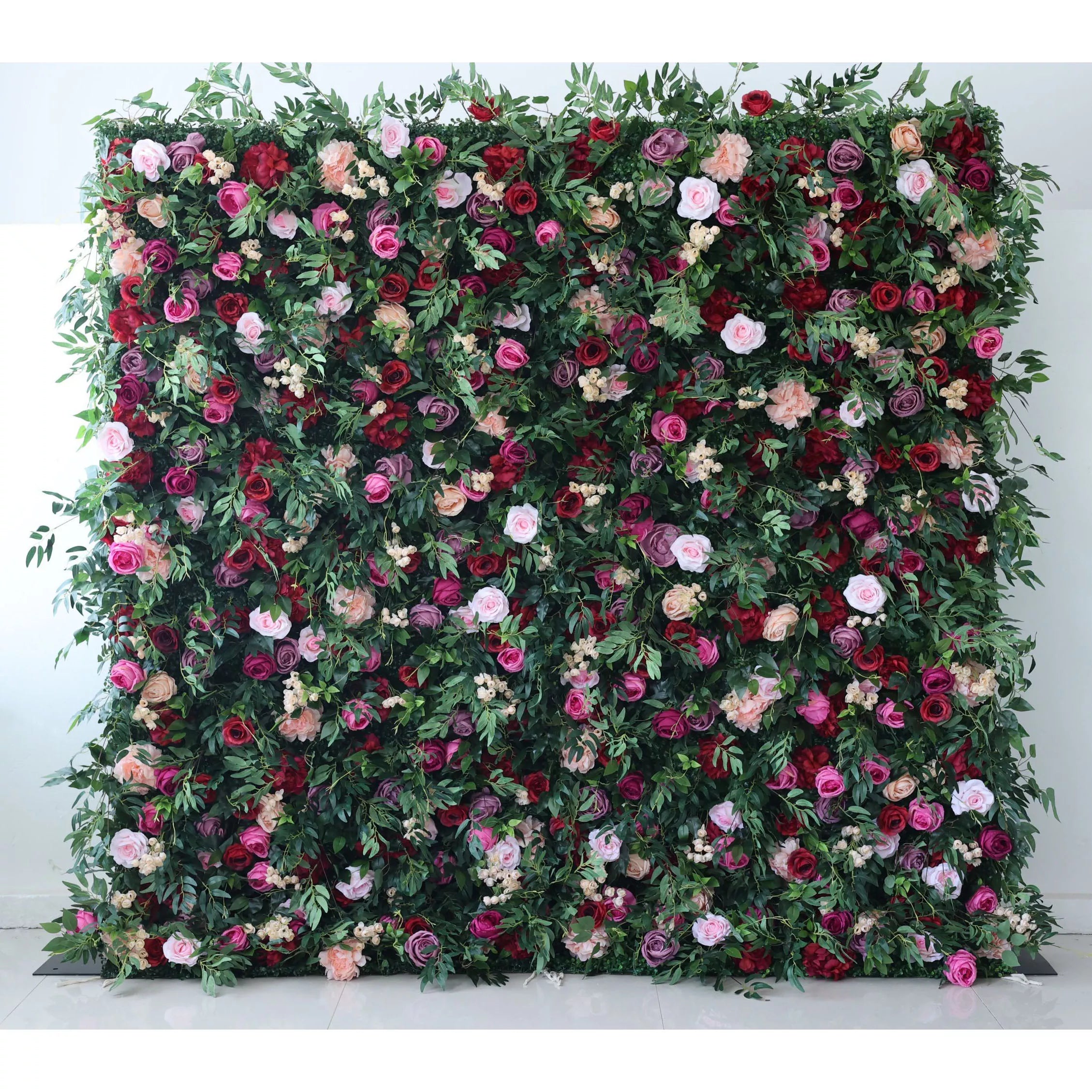 A large artificial flower wall showcases a vibrant mix of roses in pink, red, purple, and peach, interspersed with green foliage and small white blooms. It creates a lush, romantic backdrop, ideal for events or decorative spaces, exuding natural beauty and elegance.