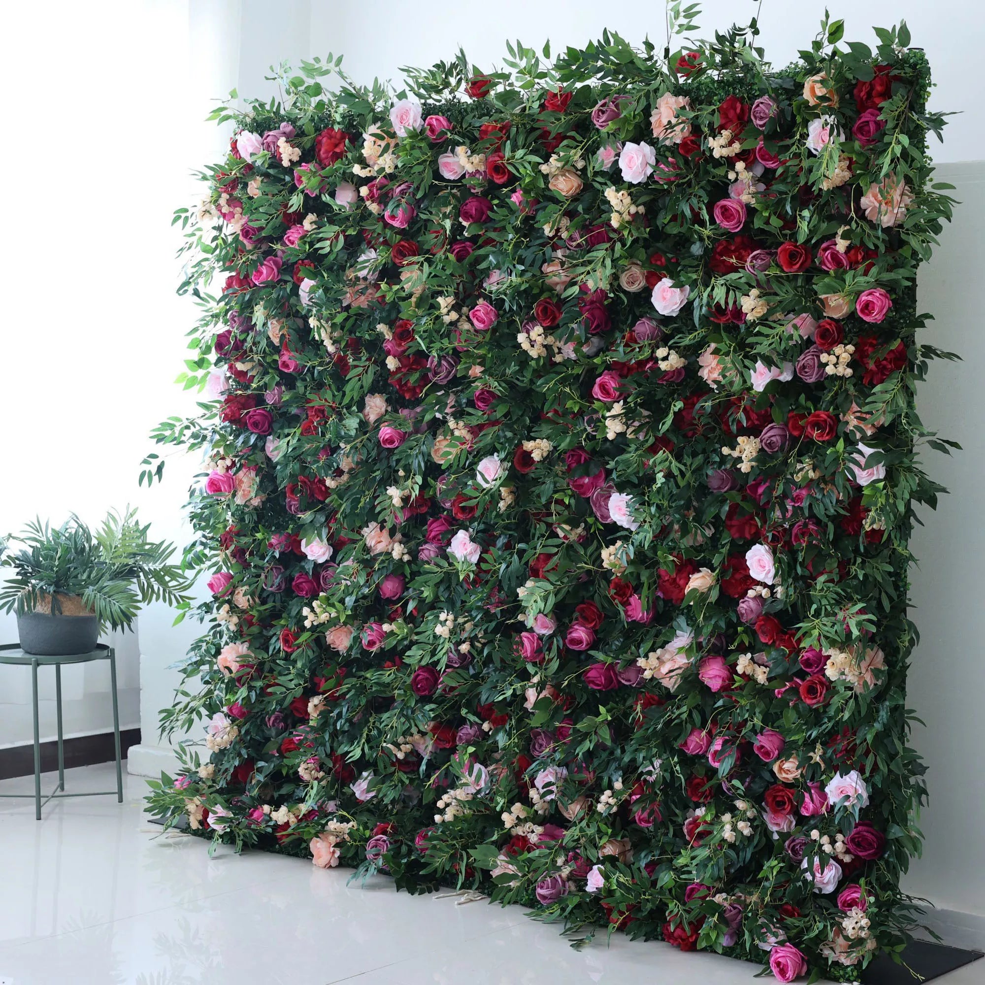 A large artificial flower wall bursts with vibrant blooms—pink, red, purple roses, plus small white flowers—all nestled in lush green foliage. It creates a lush, romantic backdrop, perfect for events or decorative spaces, adding a touch of natural beauty indoors.
