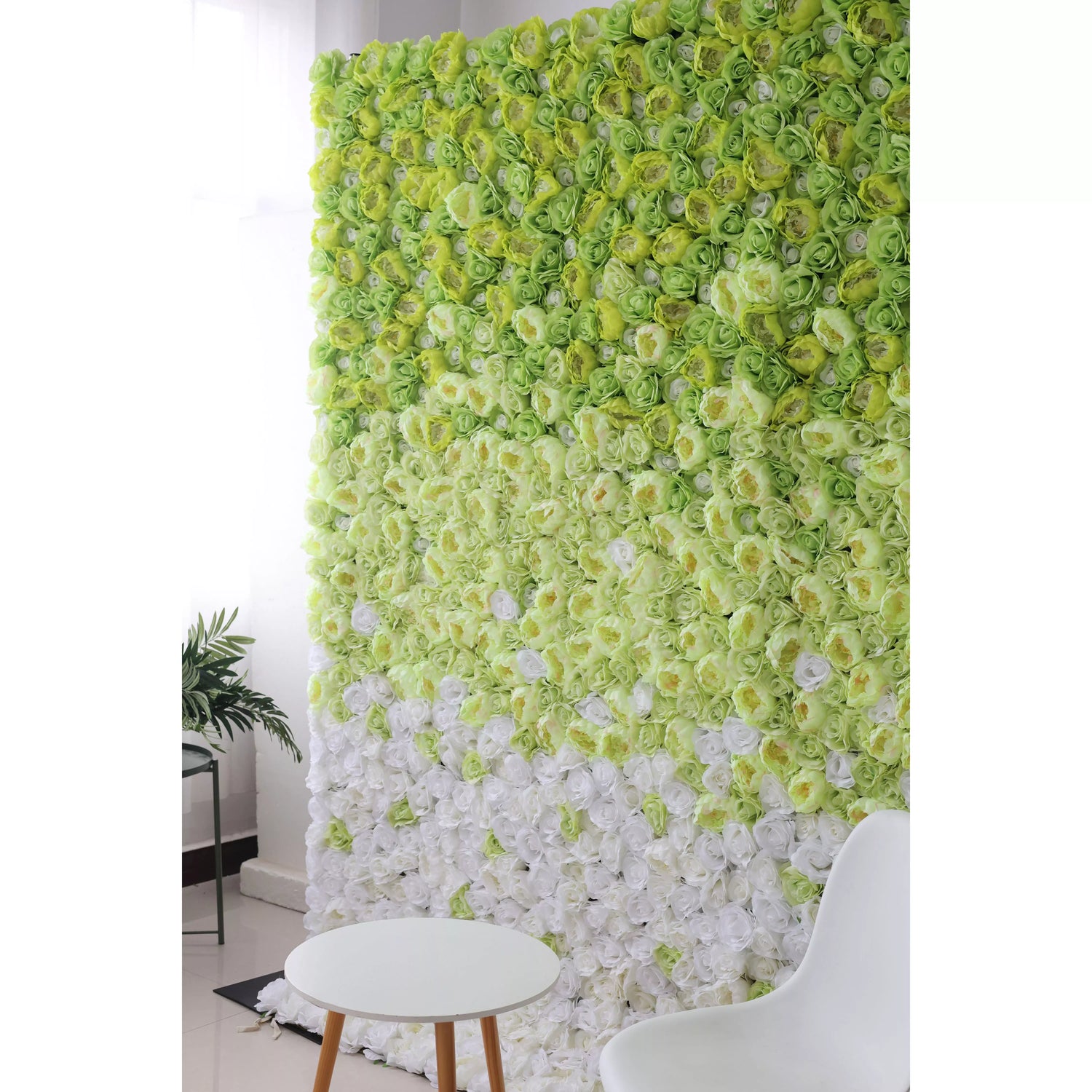 This flower wall showcases a stunning gradient of artificial roses, transitioning from vibrant green at the top to pure white at the bottom. The dense arrangement creates a lush, textured backdrop. Complemented by a minimalist white table and chair, it exudes a fresh, elegant charm, perfect for photo ops or event decor.