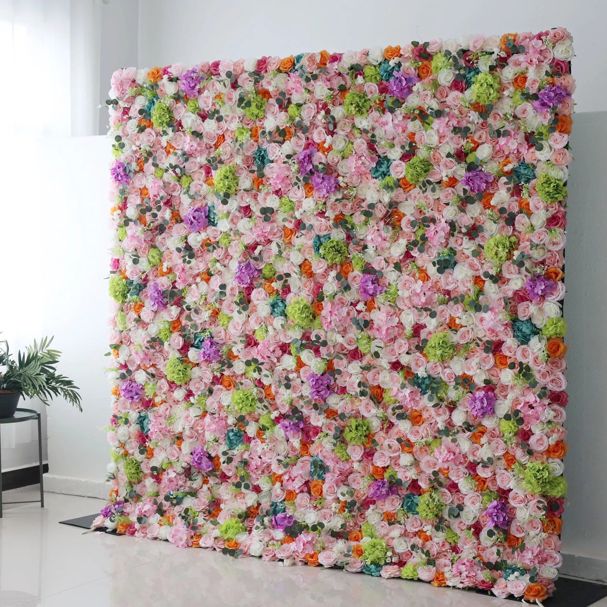 This is a vibrant, multi-colored artificial flower wall backdrop from **Valar Flowers**, bursting with a lush mix of blooms in pink, red, orange, purple, white, and green. Set indoors against a white wall, it creates a lively, cheerful, and eye-catching aesthetic ideal for weddings, photo shoots, or special events.