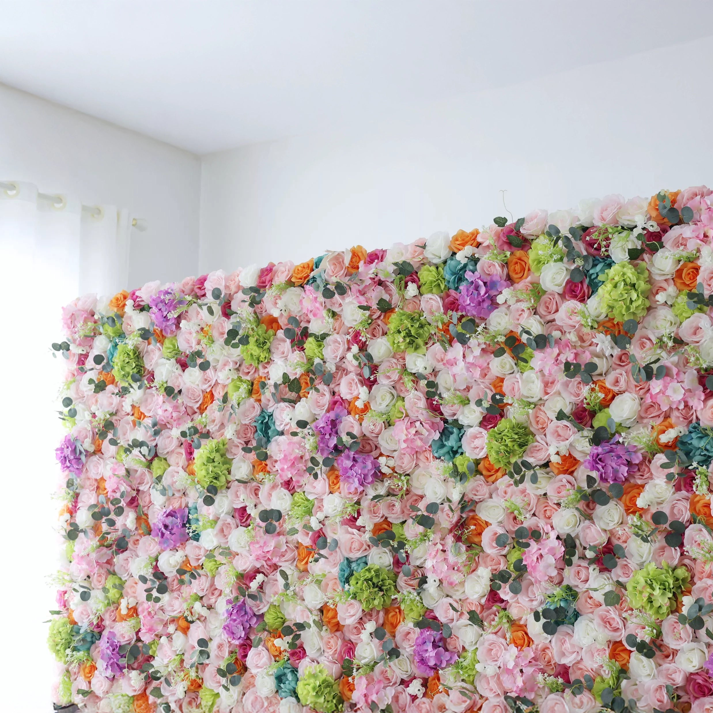 This is a close-up of a vibrant, multi-colored artificial flower wall from **Valar Flowers**, densely packed with a lush mix of blooms in soft pink, white, orange, purple, and turquoise, complemented by green hydrangeas and eucalyptus leaves. Set indoors against a white wall, it creates a lively, romantic, and eye-catching aesthetic ideal for wedding decor, photo backdrops, or special event styling.