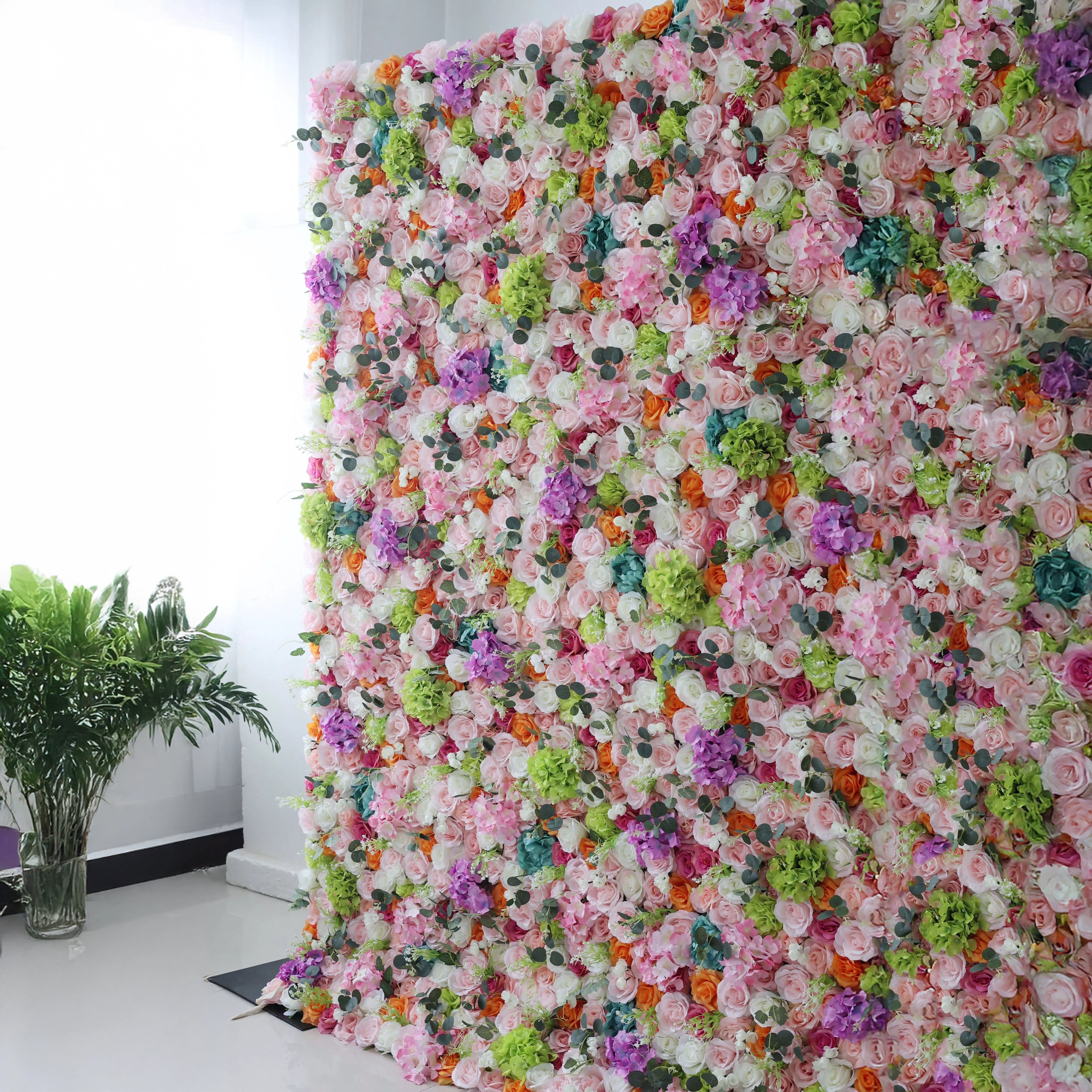 This is a vibrant, multi-colored artificial flower wall backdrop from **Valar Flowers**, densely packed with a colorful lush mix of blooms in soft pink, white, orange, purple, and turquoise, complemented by green hydrangeas and eucalyptus leaves. Set indoors against a white wall, it creates a lively, romantic, and eye-catching aesthetic ideal for wedding decor, photo backdrops, or special event styling.