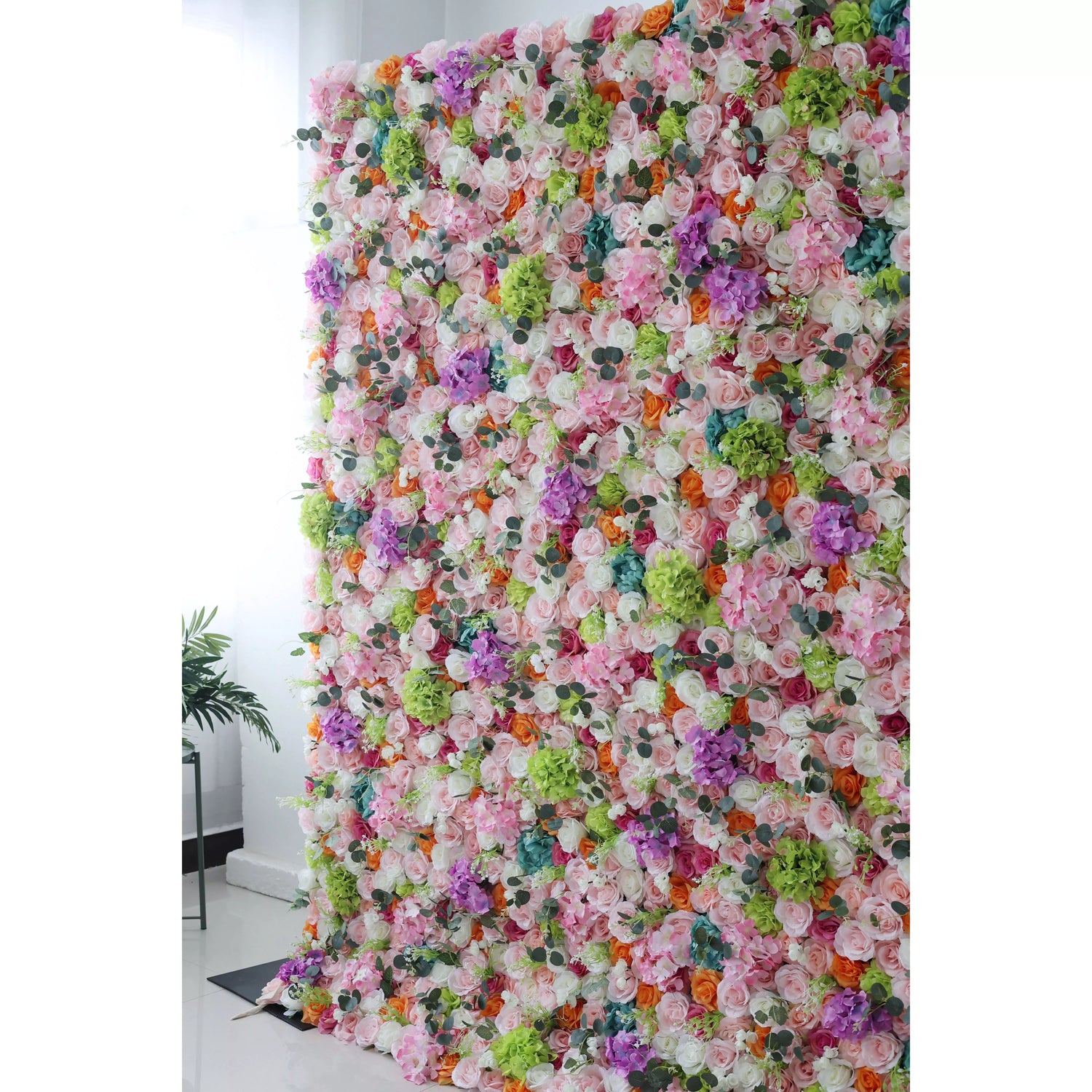 This flower wall is a vibrant, dense arrangement of artificial flowers. It features a diverse color palette—white, pink, purple, orange, green, and blue—with various blooms like roses and hydrangeas. Ideal for events, it creates a lively, celebratory atmosphere, adding charm to backdrops for photos or ceremonies.