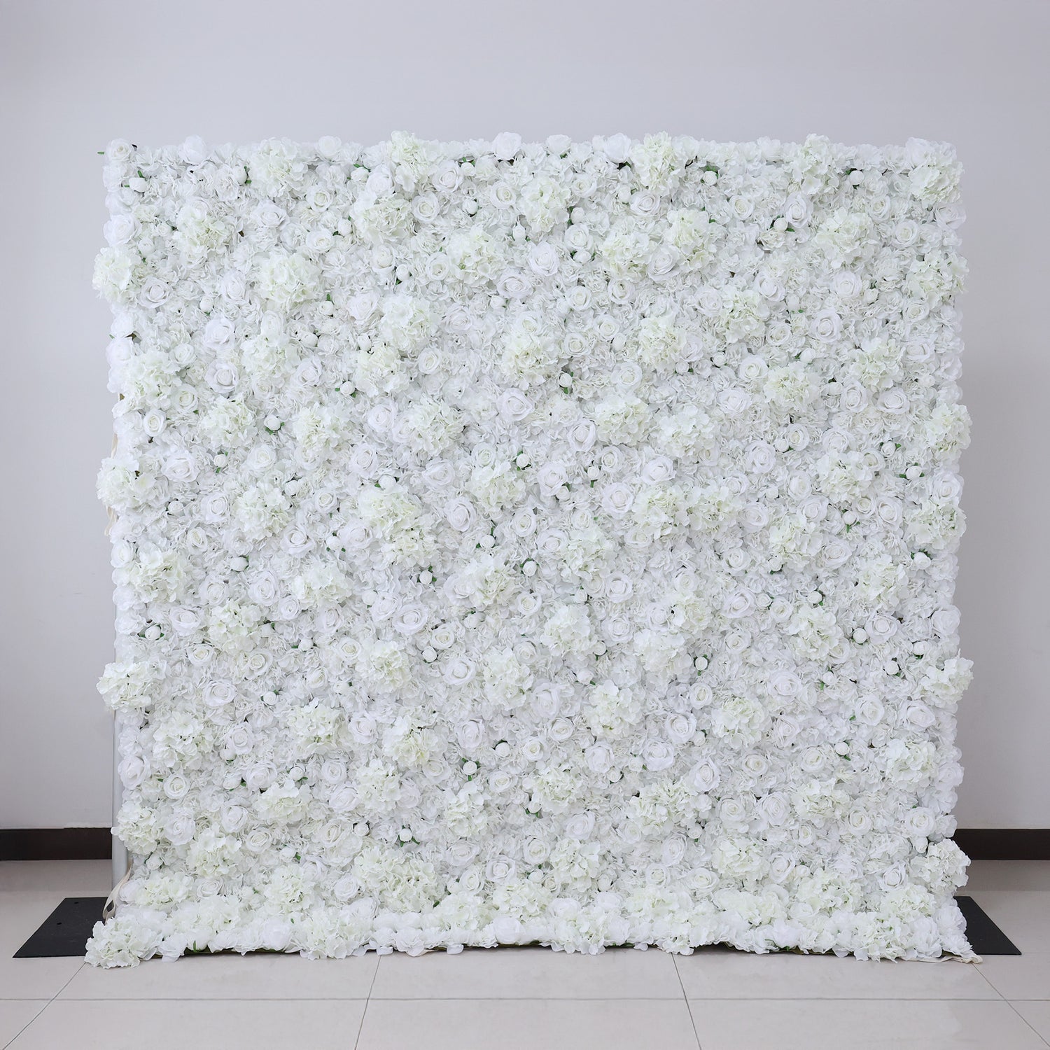 A large, dense wall of artificial white flowers fills the frame. Various white blooms, including roses and other floral types, create a lush, continuous expanse. Set against a plain light - colored wall with a tiled floor, it exudes elegance and purity, ideal for events like weddings or photo backdrops.