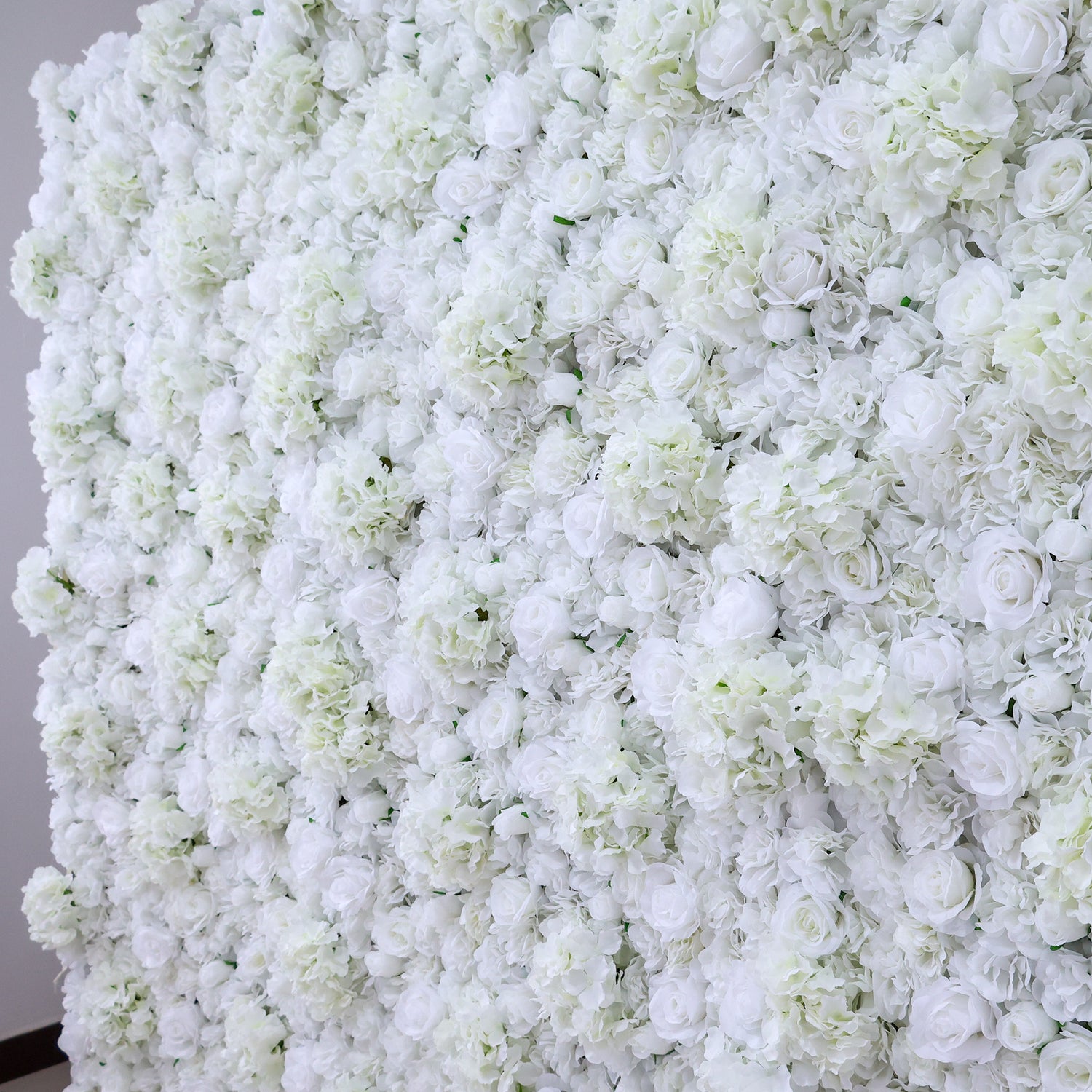 A dense wall of artificial white flowers, featuring various blooms like roses and hydrangeas, creates a lush, elegant display. The pure white hues and abundant arrangement give off a romantic, sophisticated vibe, perfect for events such as weddings, bridal showers, or upscale parties needing a beautiful backdrop.