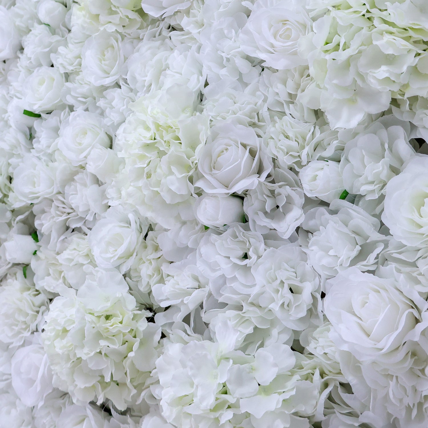 A dense wall of artificial white flowers, featuring various blooms like roses and hydrangeas, creates a lush, elegant display. The pure white hues and abundant arrangement give off a romantic, sophisticated vibe, perfect for events such as weddings, bridal showers, or upscale parties needing a beautiful backdrop.