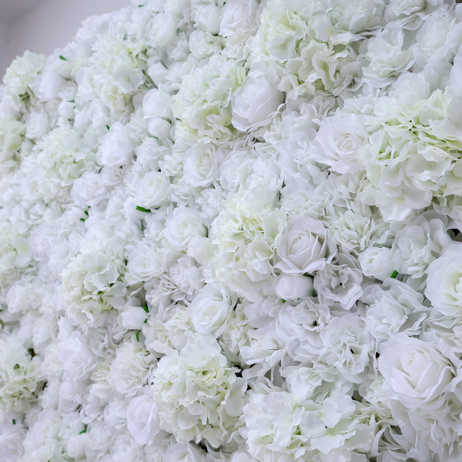A dense wall of artificial white flowers, featuring various blooms like roses and hydrangeas, creates a lush, elegant display. The pure white hues and abundant arrangement give off a romantic, sophisticated vibe, perfect for events such as weddings, bridal showers, or upscale parties needing a beautiful backdrop.