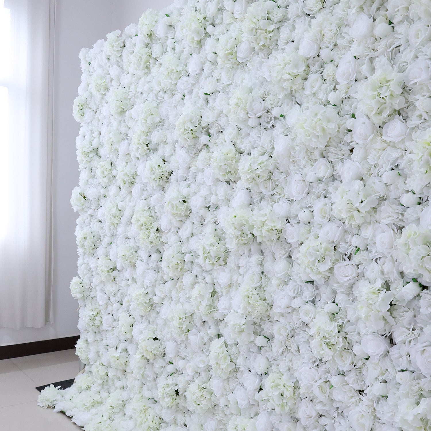 A large, dense wall of artificial white flowers fills the frame, showcasing a lush arrangement of various blooms like roses and hydrangeas. Set in a room with light - colored walls, a tiled floor, and a window with sheer curtains allowing soft light to filter in, it exudes an elegant and romantic vibe, perfect for events such as weddings, bridal showers, or upscale gatherings needing a sophisticated backdrop.