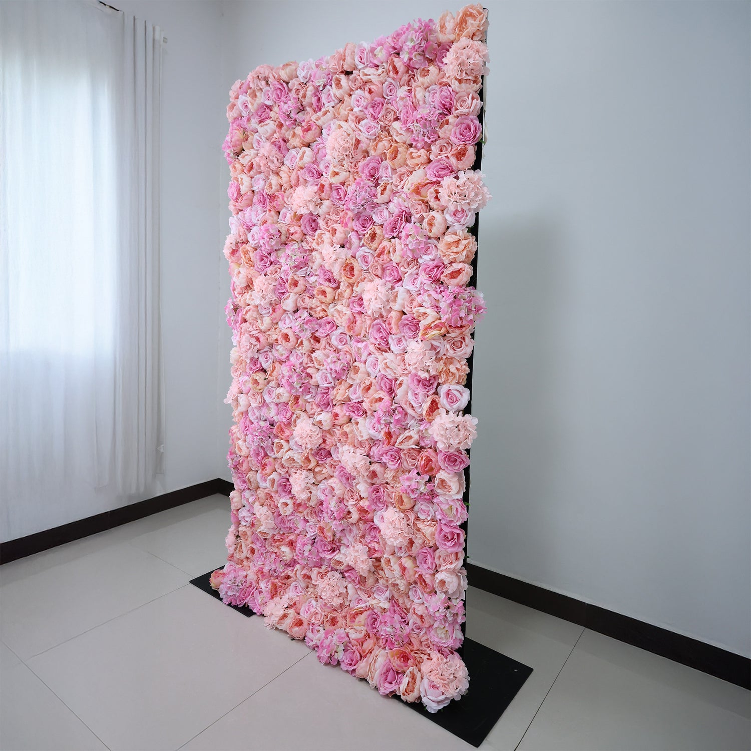 This image showcases a stunning pink floral backdrop by ValarFlowers, set indoors. It features a dense arrangement of artificial flowers in various shades of pink, creating a lush, textured display. Natural light from a window enhances the scene, exuding elegance and sophistication, perfect for adding a luxurious, nature-inspired element to events or interiors.