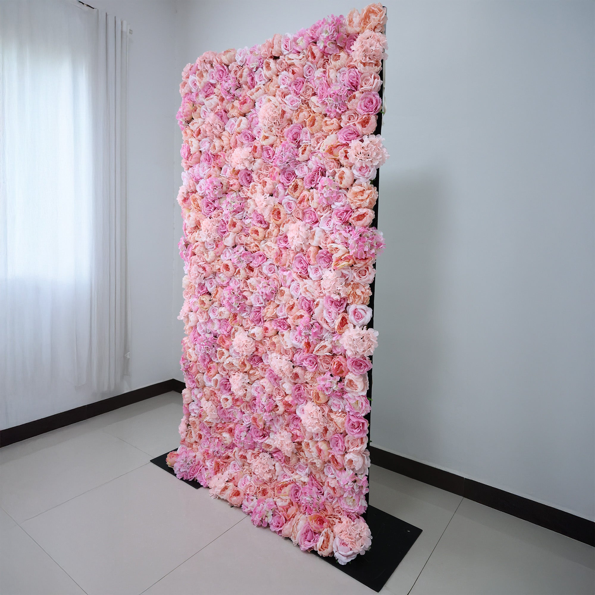 This image showcases a stunning pink floral backdrop by ValarFlowers, set indoors. It features a dense arrangement of artificial flowers in various shades of pink, creating a lush, textured display. Natural light from a window enhances the scene, exuding elegance and sophistication, perfect for adding a luxurious, nature-inspired element to events or interiors.