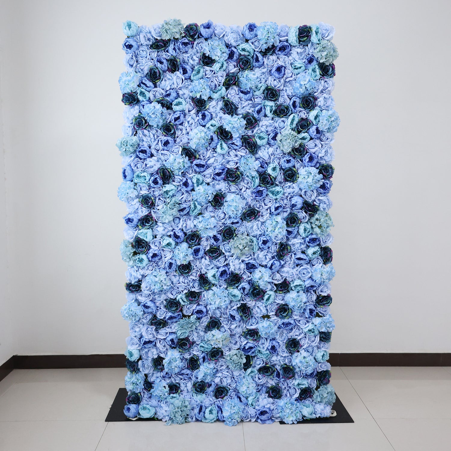 This image showcases a stunning floral backdrop by ValarFlowers, measuring 4x8ft (1.2x2.4m), set indoors on a glossy floor. It features a dense arrangement of artificial flowers in various shades of blue—from light sky blue to deep navy—including roses and hydrangeas, creating a lush, textured, and visually captivating display. The harmonious blend of hues and realistic floral details exude elegance and sophistication, making it perfect for adding a luxurious, nature-inspired element to events or interiors.