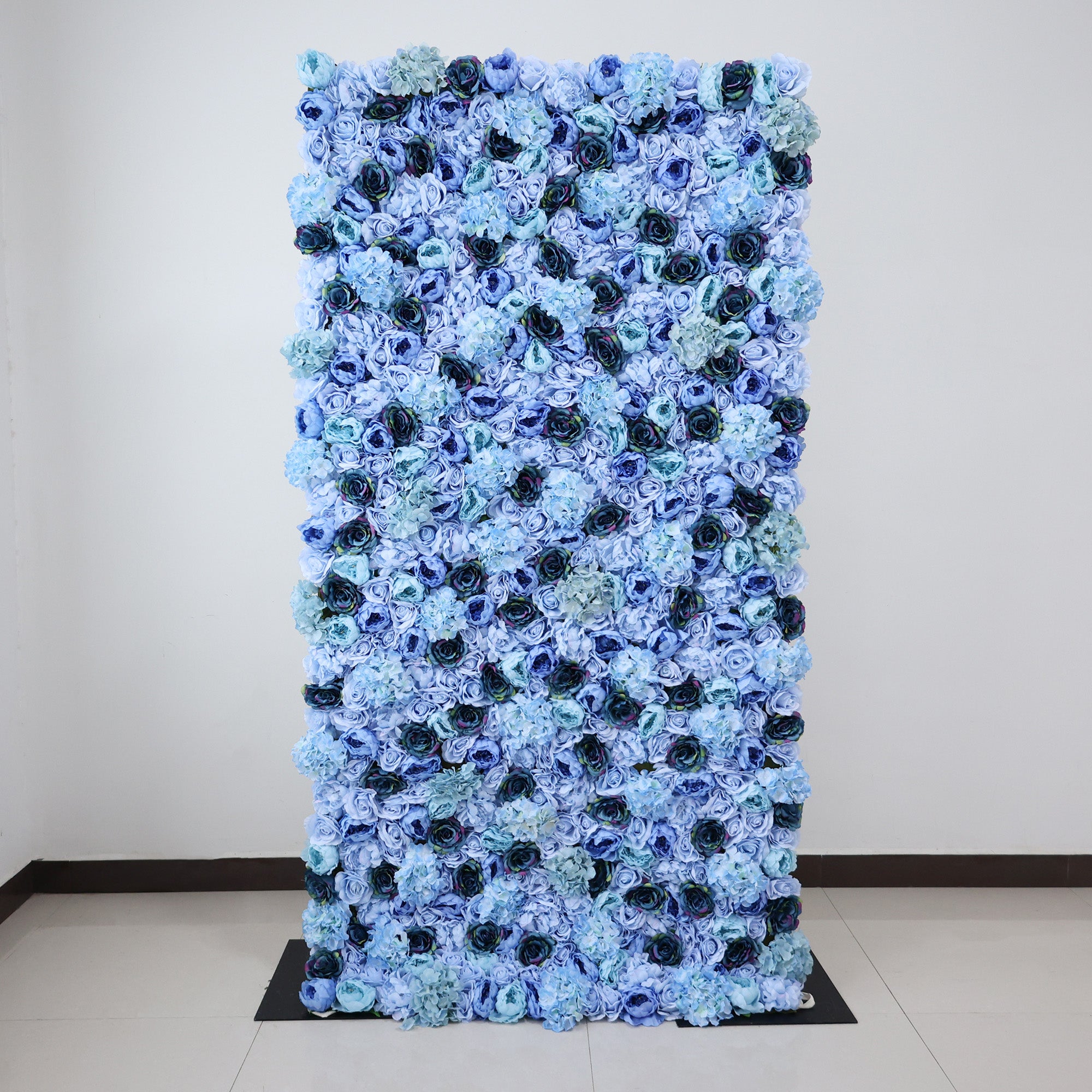 This image showcases a stunning floral backdrop by ValarFlowers, measuring 4x8ft (1.2x2.4m), set indoors on a glossy floor. It features a dense arrangement of artificial flowers in various shades of blue—from light sky blue to deep navy—including roses and hydrangeas, creating a lush, textured, and visually captivating display. The harmonious blend of hues and realistic floral details exude elegance and sophistication, making it perfect for adding a luxurious, nature-inspired element to events or interiors.