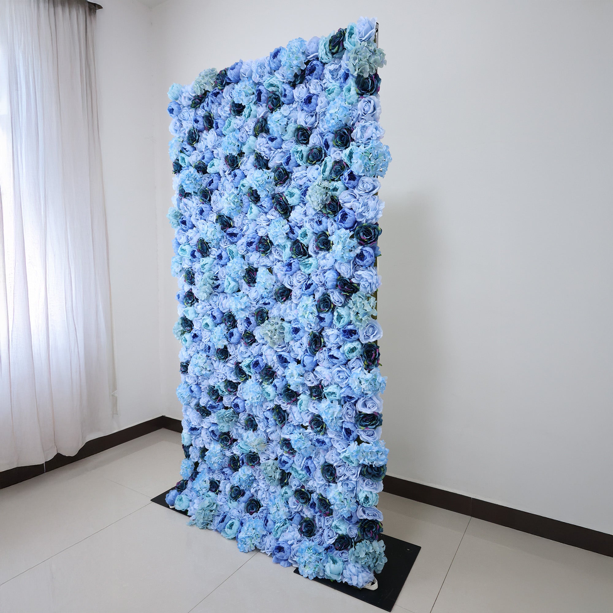 This image showcases a stunning blue floral backdrop by ValarFlowers, set indoors. It features dense artificial flowers in various shades of blue, including roses and hydrangeas, creating a lush, textured display. The elegant design, complemented by natural light from a window, exudes sophistication, ideal for adding a luxurious, nature-inspired element to events or interiors.