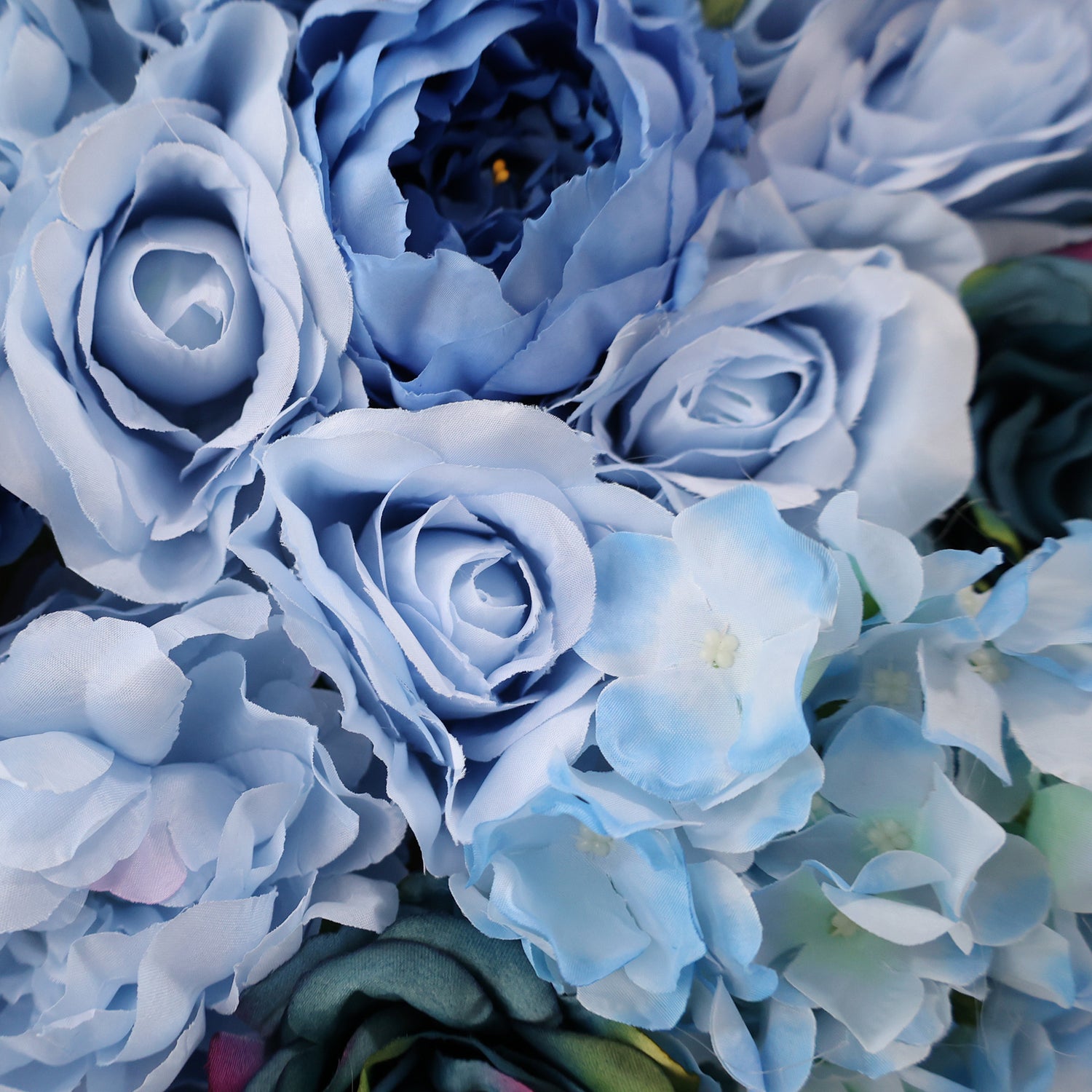 This image showcases a dense arrangement of artificial flowers in various shades of blue, including roses and hydrangeas. The soft, textured petals in hues from light sky blue to deep navy create a lush, elegant display, ideal for a luxurious, nature-inspired backdrop, perfect for adding sophistication to events or interiors.