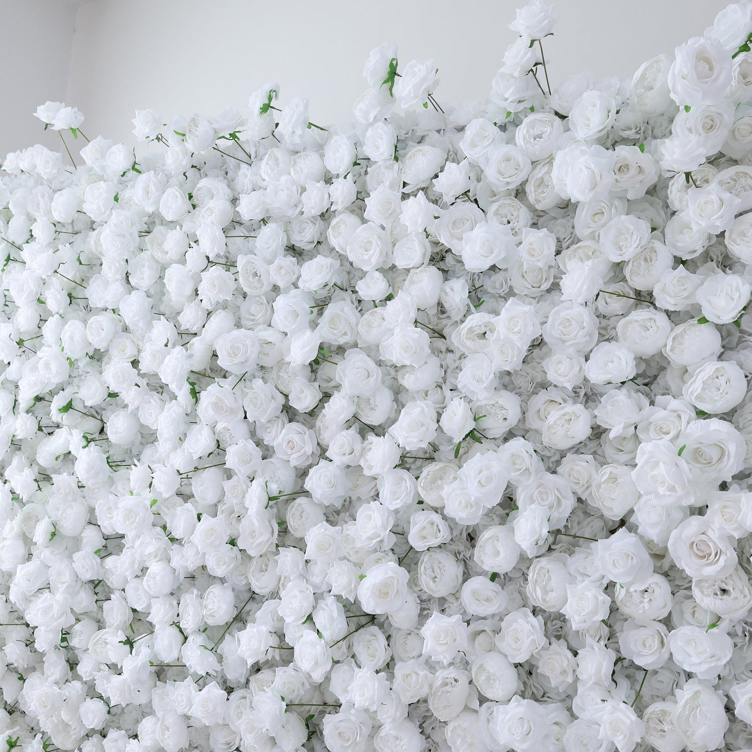 This image presents a dense arrangement of white artificial roses, creating a lush and voluminous floral display. The flowers are tightly packed, with their soft, creamy petals and subtle green stems contributing to a sense of purity and elegance. Set indoors against a plain white wall, the scene exudes a serene and sophisticated atmosphere, ideal for decorative purposes in events or as a striking interior backdrop, evoking feelings of natural beauty and refinement.