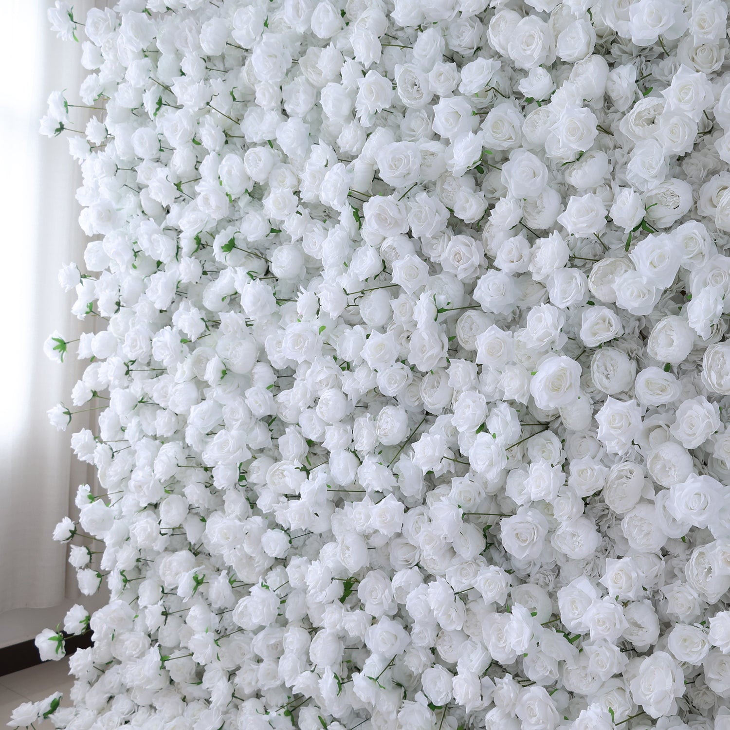 This image presents a dense arrangement of white artificial roses, creating a lush and voluminous floral display. The flowers are tightly packed, with their soft, creamy petals and subtle green stems contributing to a sense of purity and elegance. Set indoors, likely near a window with light filtering through, the scene exudes a serene and sophisticated atmosphere, ideal for decorative purposes in events or as a striking interior backdrop, evoking feelings of natural beauty and refinement.