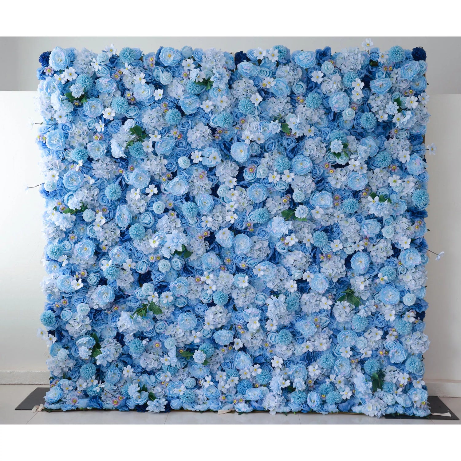 A large artificial flower wall features a lush arrangement of blue blooms, including roses and hydrangeas, interspersed with tiny white flowers and green foliage. The varying shades of blue create a harmonious, serene backdrop, perfect for adding an elegant, calming touch to events or decor.