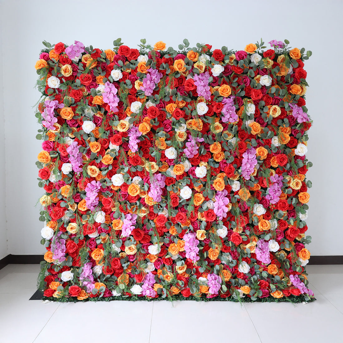 This image showcases a vibrant floral wall by Valar Flowers, set indoors against a plain white wall and tiled floor. It features a dense arrangement of artificial flowers in a rich palette of red, orange, pink, and white, creating a lively and eye-catching display. The backdrop is perfect for events, with the bold hues and lush texture adding a touch of energy and elegance, ideal for enhancing the ambiance of weddings or photo shoots.