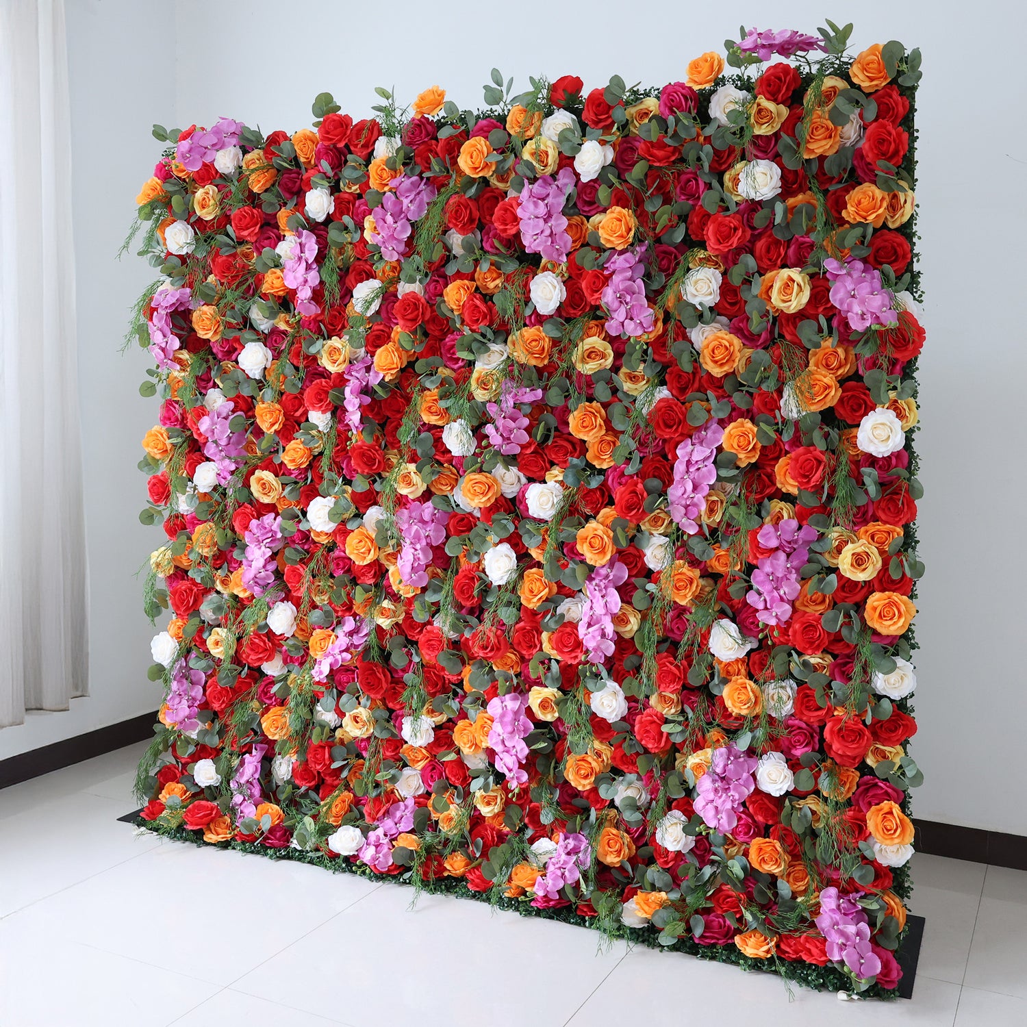 This image showcases a vibrant floral wall by Valar Flowers, set indoors against a plain white wall and tiled floor. It features a dense arrangement of artificial flowers in a rich palette of red, orange, pink, and white, creating a lively and eye-catching display. The backdrop is perfect for events, with the bold hues and lush texture adding a touch of energy and elegance, ideal for enhancing the ambiance of weddings or photo shoots.