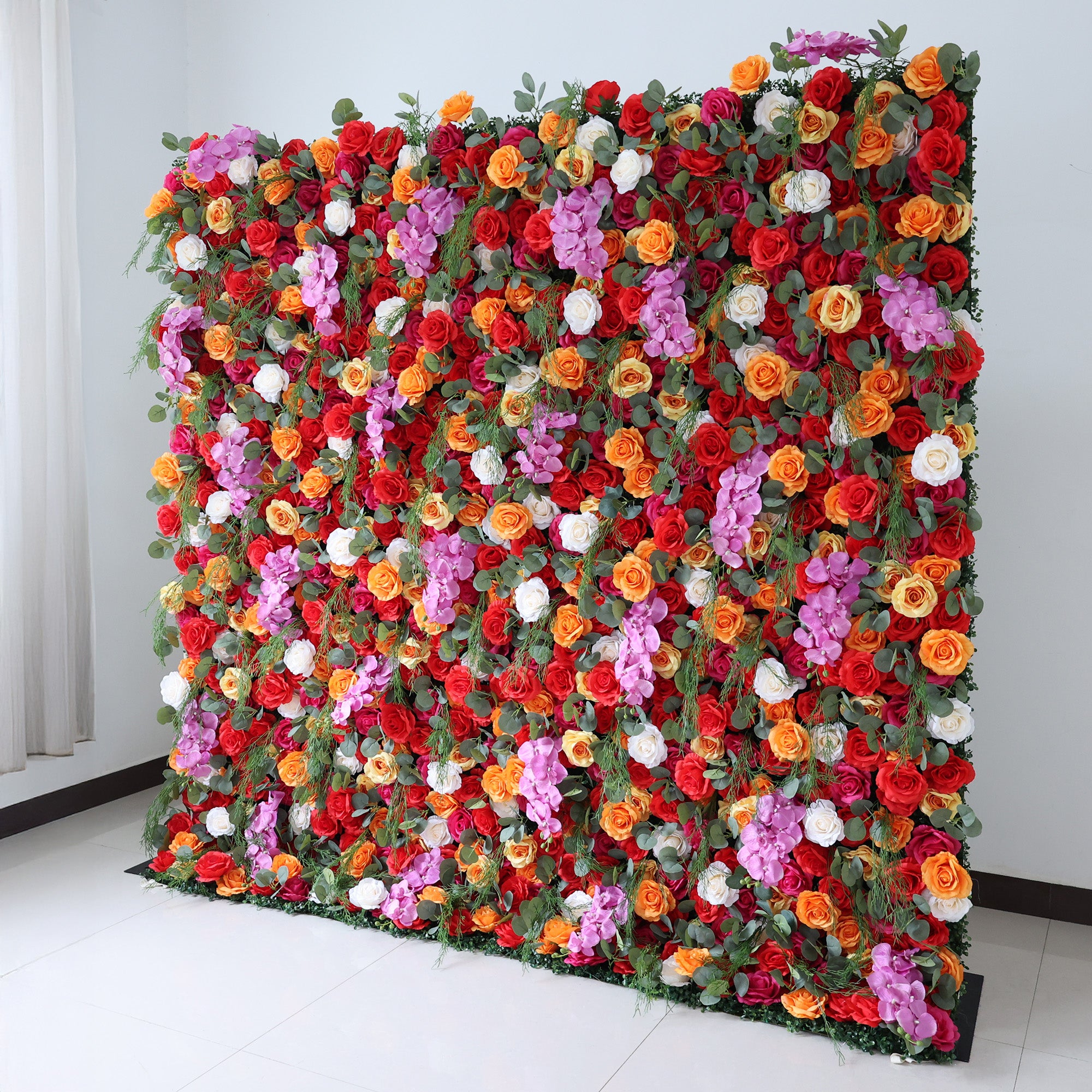 This image showcases a vibrant floral wall by Valar Flowers, set indoors against a plain white wall and tiled floor. It features a dense arrangement of artificial flowers in a rich palette of red, orange, pink, and white, creating a lively and eye-catching display. The backdrop is perfect for events, with the bold hues and lush texture adding a touch of energy and elegance, ideal for enhancing the ambiance of weddings or photo shoots.