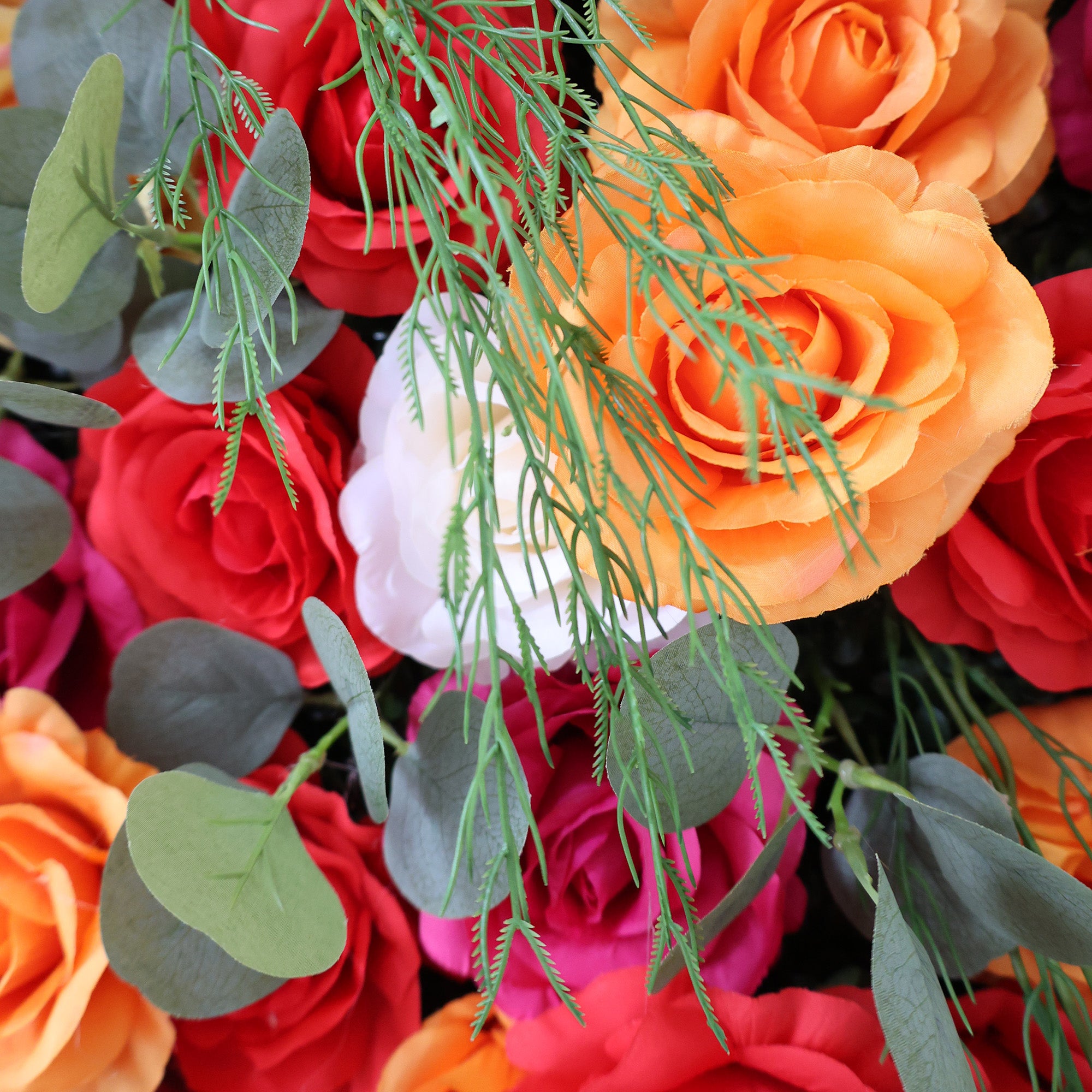 This image showcases a vibrant floral arrangement by Valar Flowers. It features a dense cluster of artificial flowers in a rich palette of red, orange, pink, and white, including roses and other complementary blooms, interspersed with lush green foliage. The flowers are intricately arranged to create a lush, textured, and visually striking display. Perfect for adding a touch of energy and elegance to events or spaces, the arrangement exudes a lively and sophisticated atmosphere.