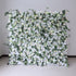 This image showcases a stunning floral wall by Valar Flowers. It features a dense arrangement of artificial flowers in soft shades of white and cream, interspersed with lush green foliage. The meticulously crafted blooms create a lush, elegant, and visually soothing display. Perfect for adding a touch of timeless beauty to events or spaces, this floral wall exudes a refined and sophisticated atmosphere.