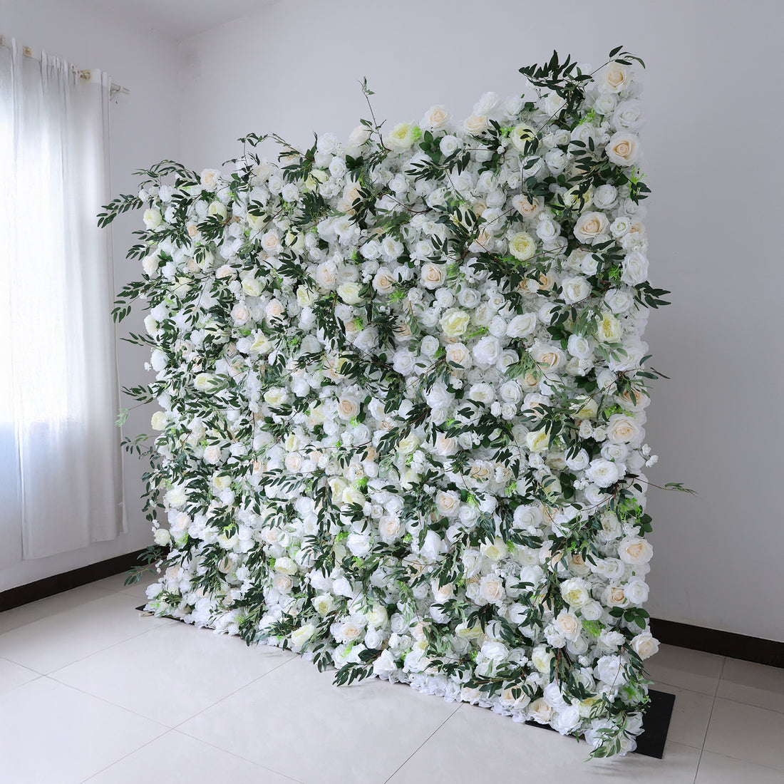 This image showcases a stunning floral wall by Valar Flowers. It features a dense arrangement of artificial flowers in soft shades of white and cream, interspersed with lush green foliage. The meticulously crafted blooms create a lush, elegant, and visually soothing display. Set indoors against a plain white wall and tiled floor, this floral wall is perfect for adding a touch of timeless beauty to events or spaces, exuding a refined and sophisticated atmosphere.